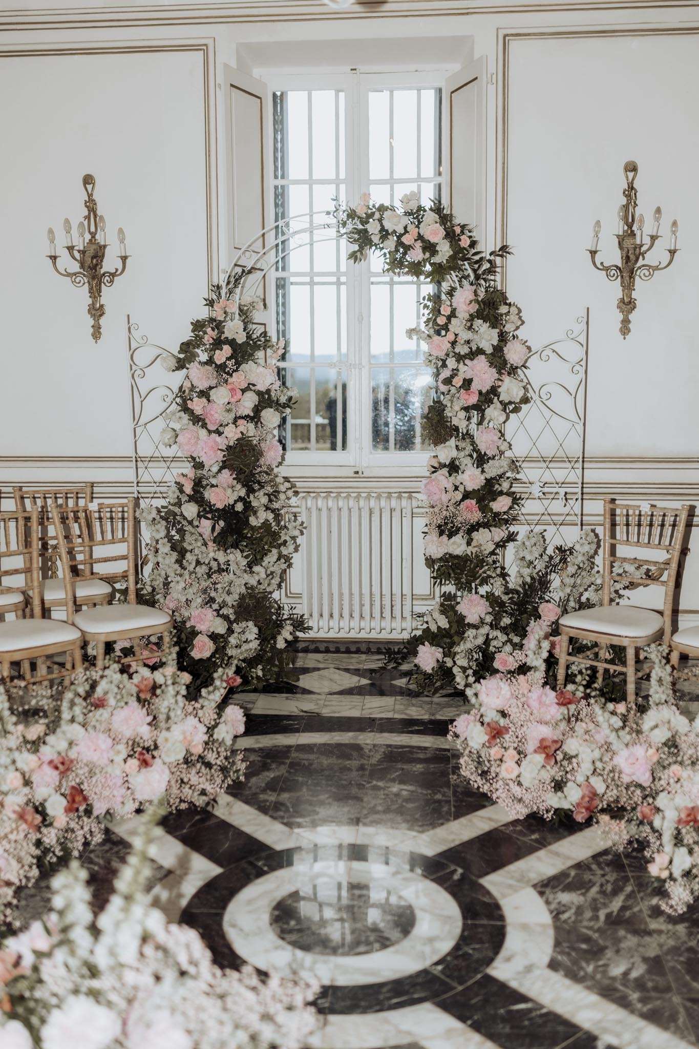 Indoor ceremony setup with circular floral arch of pink peonies and white roses, gold chiavari chairs on marble floor