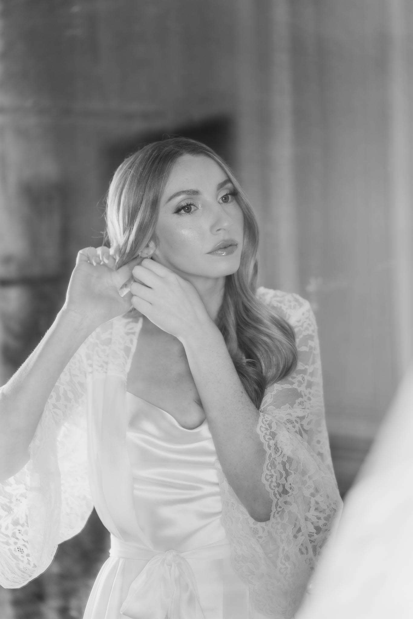 Black-and-white portrait of bride fastening earring in lace-sleeved robe over satin slip during getting ready