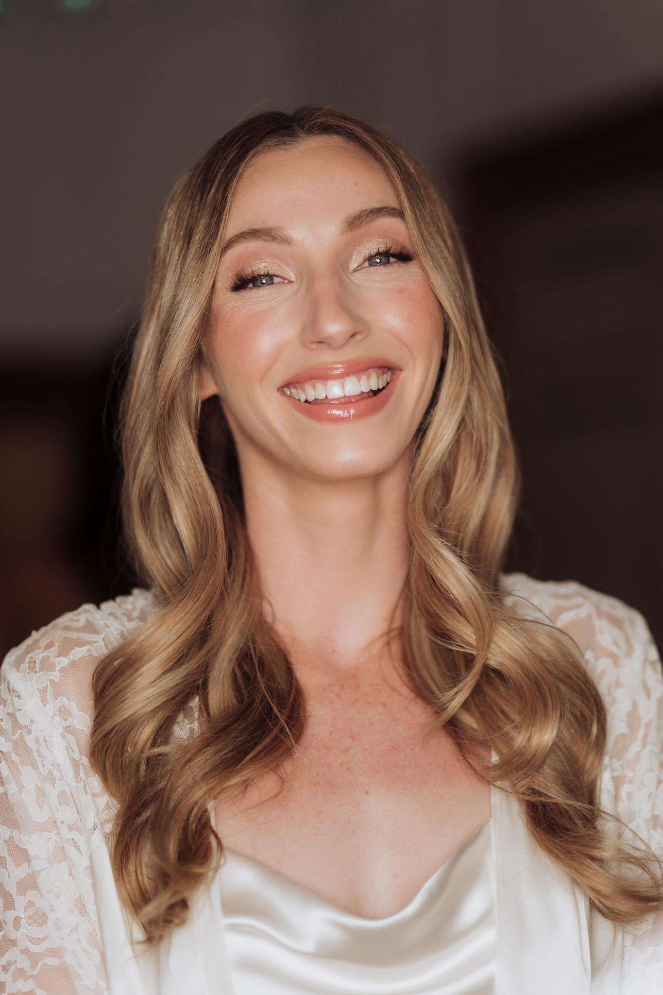 A close-up portrait of a bride during the getting-ready phase, photographed indoors against a softly blurred dark background. She wears a white satin cowl-neck slip and a white lace robe over it, with long blonde wavy hair worn loose and styled with voluminous curls. Her makeup is natural and polished, featuring a nude-pink glossy lip, subtle shimmer eyeshadow, and defined lashes. The shot is tightly framed from the shoulders up, with warm, soft lighting illuminating her face.