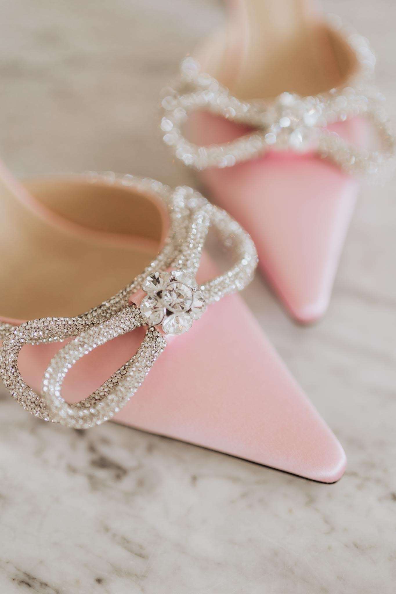 Close-up detail shot of a pair of bridal heels in pale pink satin, featuring pointed toes and crystal-encrusted bow embellishments at the vamp. Each bow is outlined in pavé rhinestones and centered with three larger faceted crystal stones, with pearl-beaded trim visible around the collar of the shoe. The shoes are placed on a white marble surface. The foreground shoe is in sharp focus while the second shoe is softly blurred in the background, creating depth. The overall styling aesthetic is feminine and ornate, consistent with a modern bridal look.