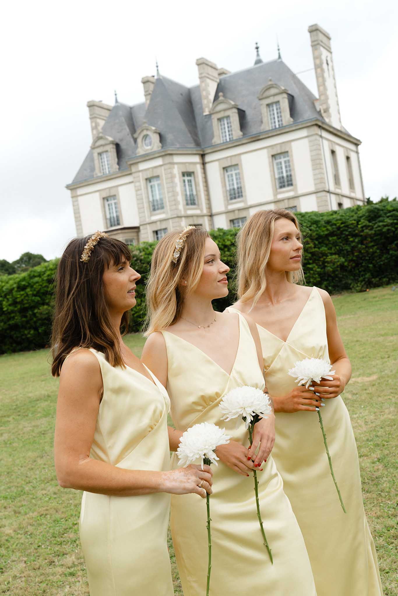 Three bridesmaids in pale yellow dresses holding white dahlias on lawn before 19th-century French chateau