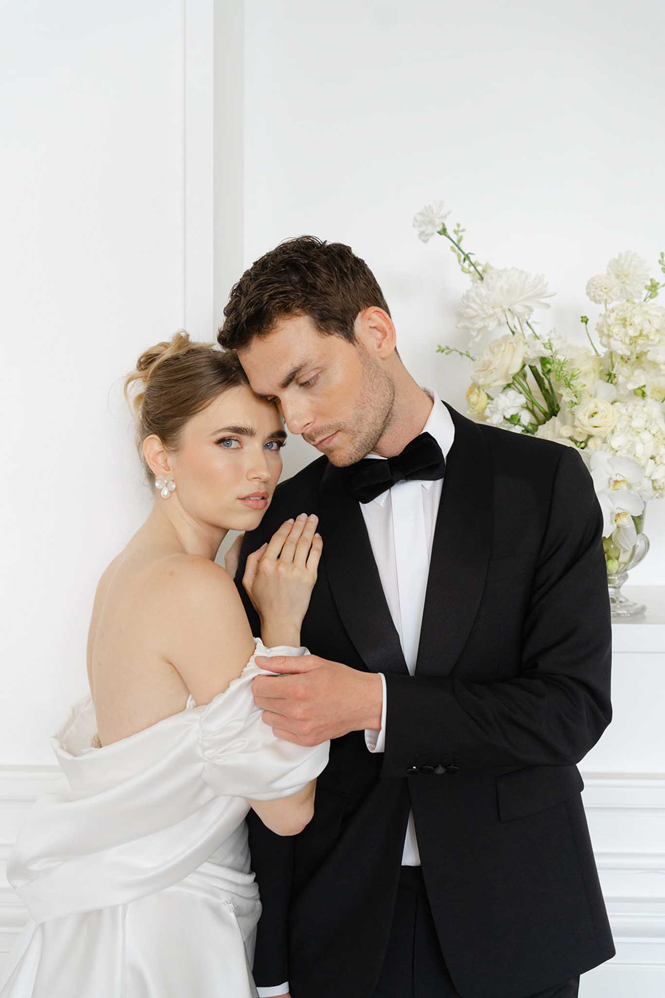 Bride in off-shoulder ivory dress and groom in black tuxedo pose indoors near white carnation arrangement