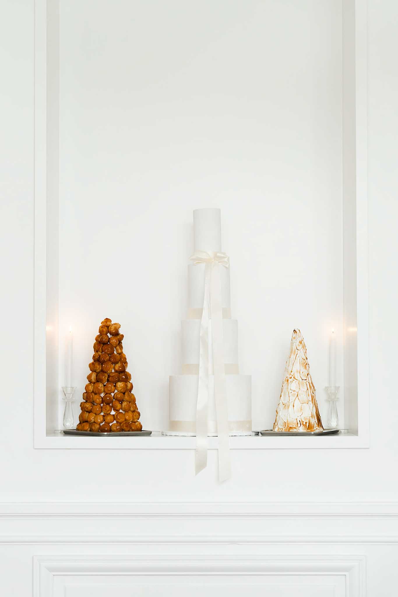 Dessert display on white mantelpiece: croquembouche, three-tiered white wedding cake and caramel dessert tower with candles