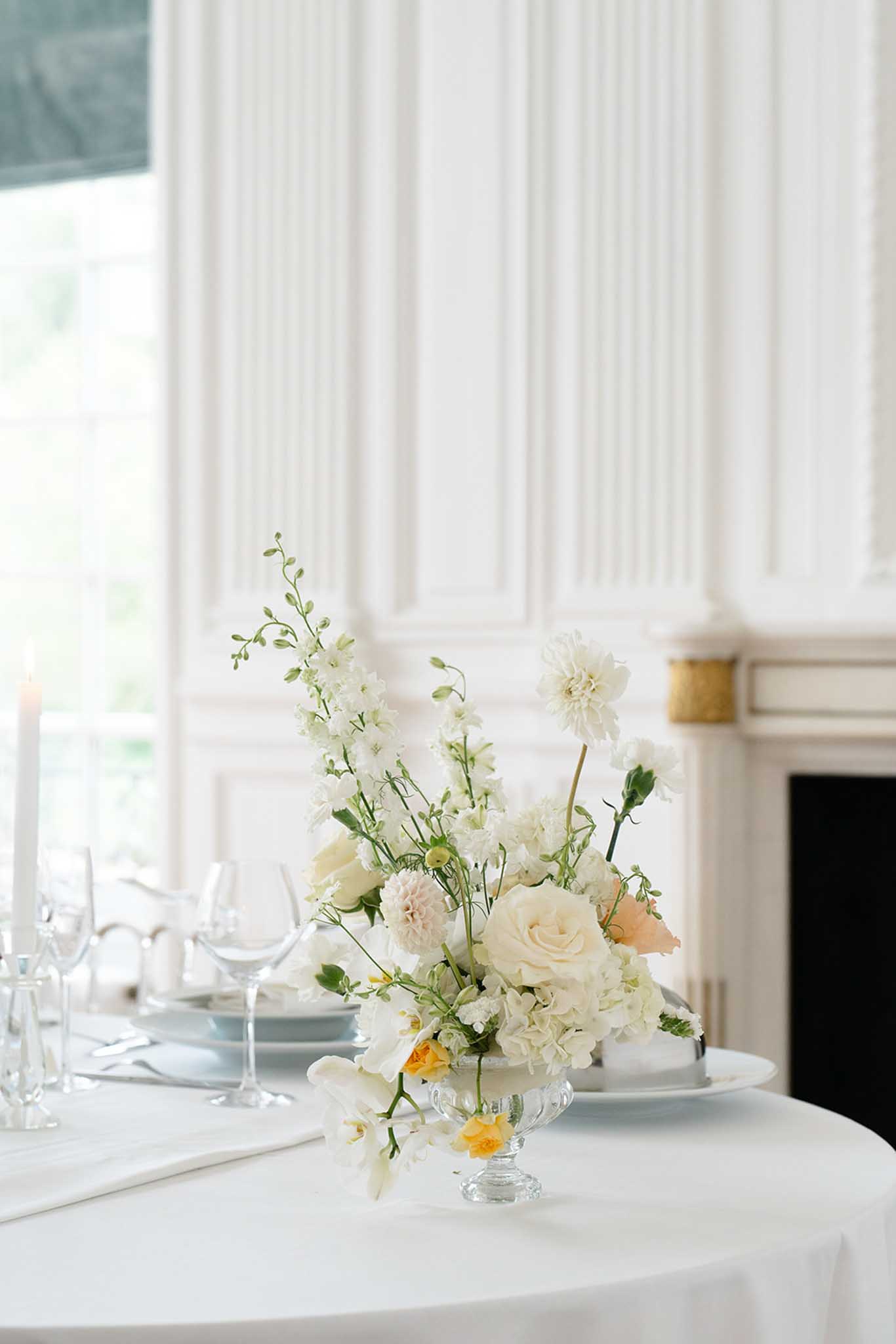 Reception table centerpiece of cream roses, white dahlias, and ranunculus in a glass vase with gold fireplace backdrop