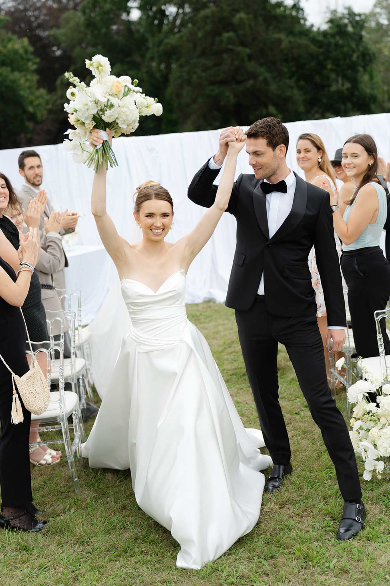 Bride raising bouquet triumphantly during recessional with groom on garden lawn as guests applaud