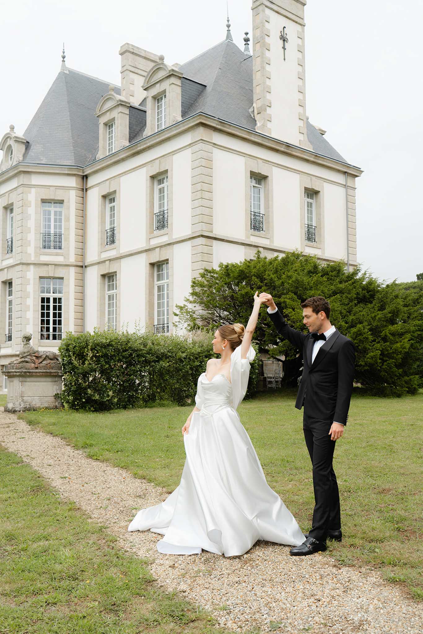 Bride and groom celebrating on gravel pathway with Mansard-roofed French chateau and hedged gardens behind