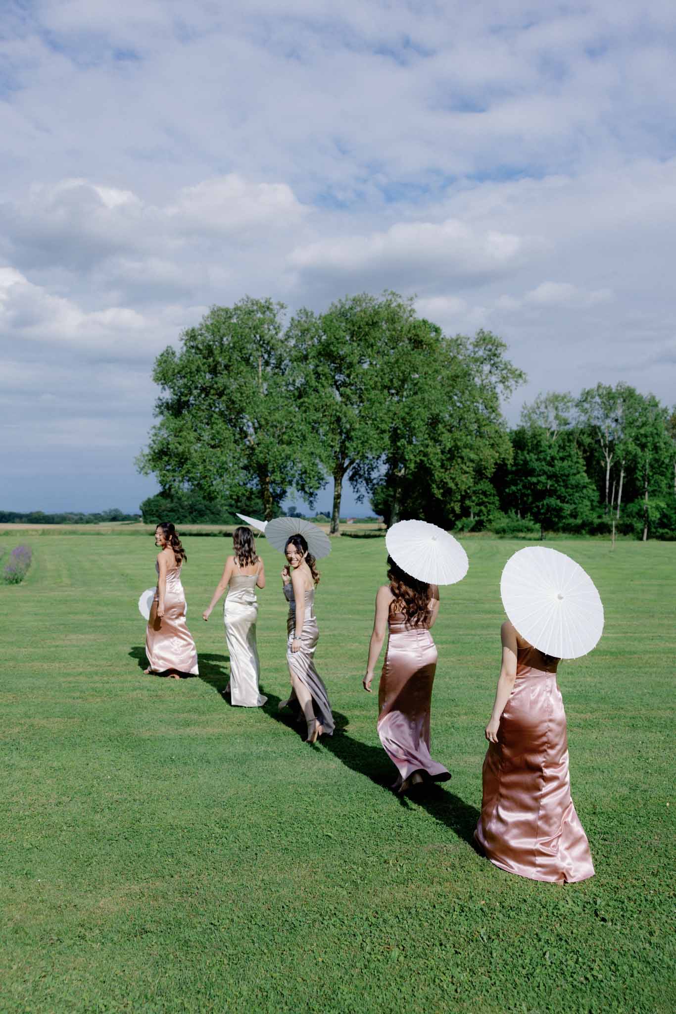 Five bridesmaids walking across lawn in floor-length satin dresses in blush pink and champagne tones