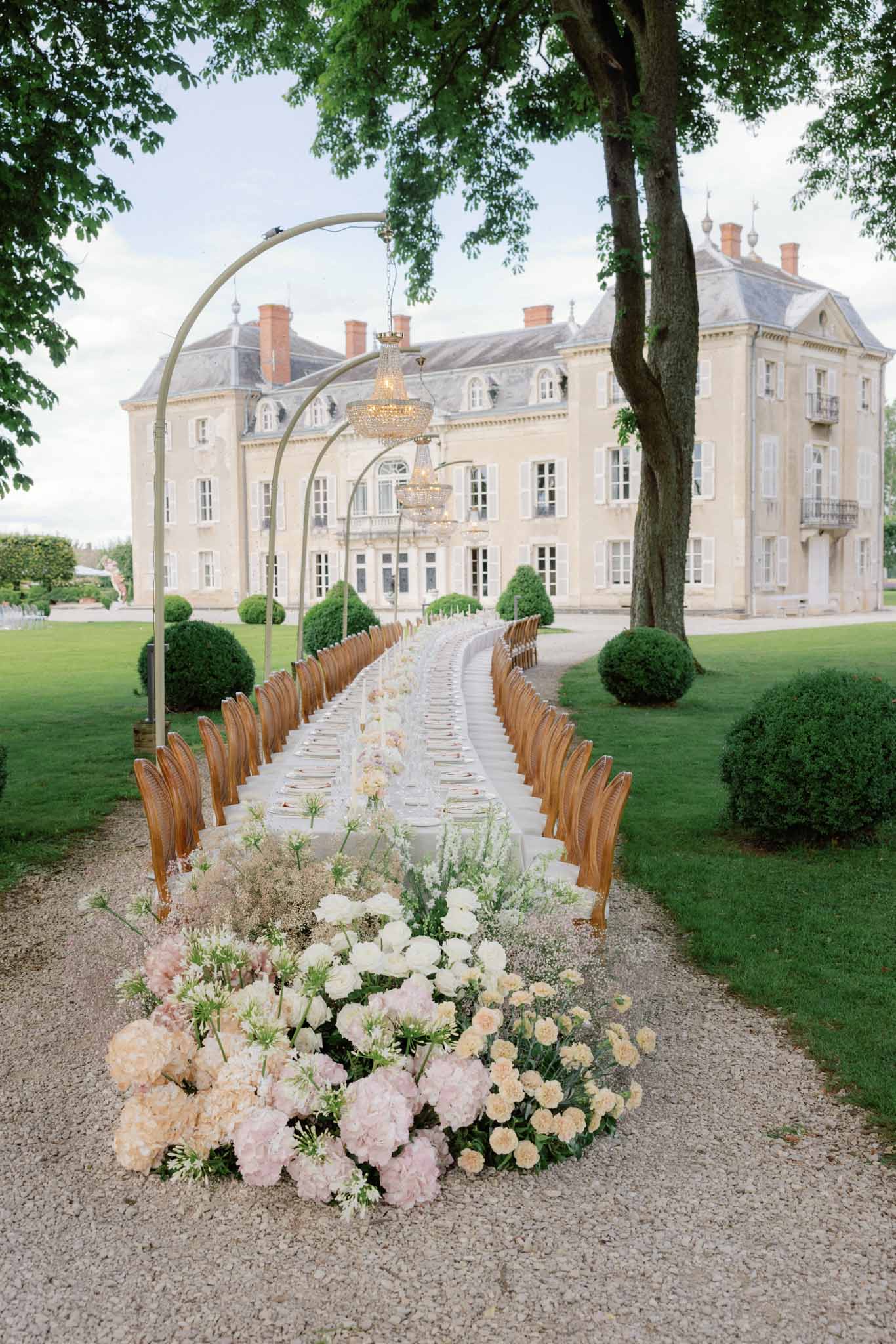 Ceremony aisle with wooden chairs and floral arrangements leading to a classical chateau entrance with chandelier arch