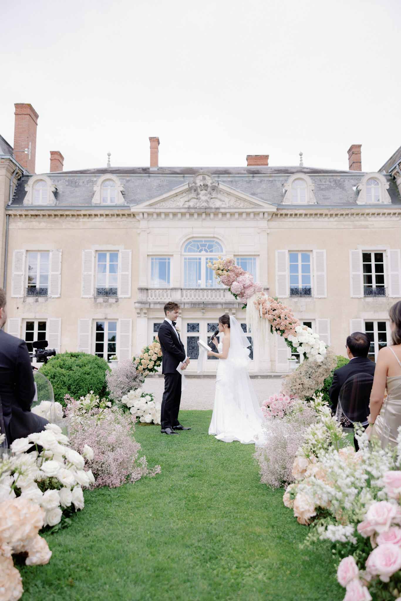 Wedding ceremony on chÃ¢teau lawn with couple beneath blush and white floral arch, guests lining aisle of rose and hydrangea clusters