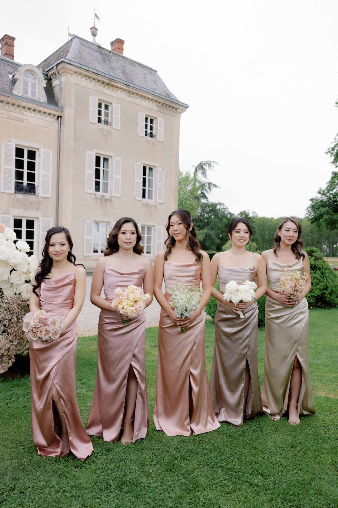 Five bridesmaids in gradient dusty rose and champagne satin gowns standing in line on the lawn of a French chÃ¢teau.