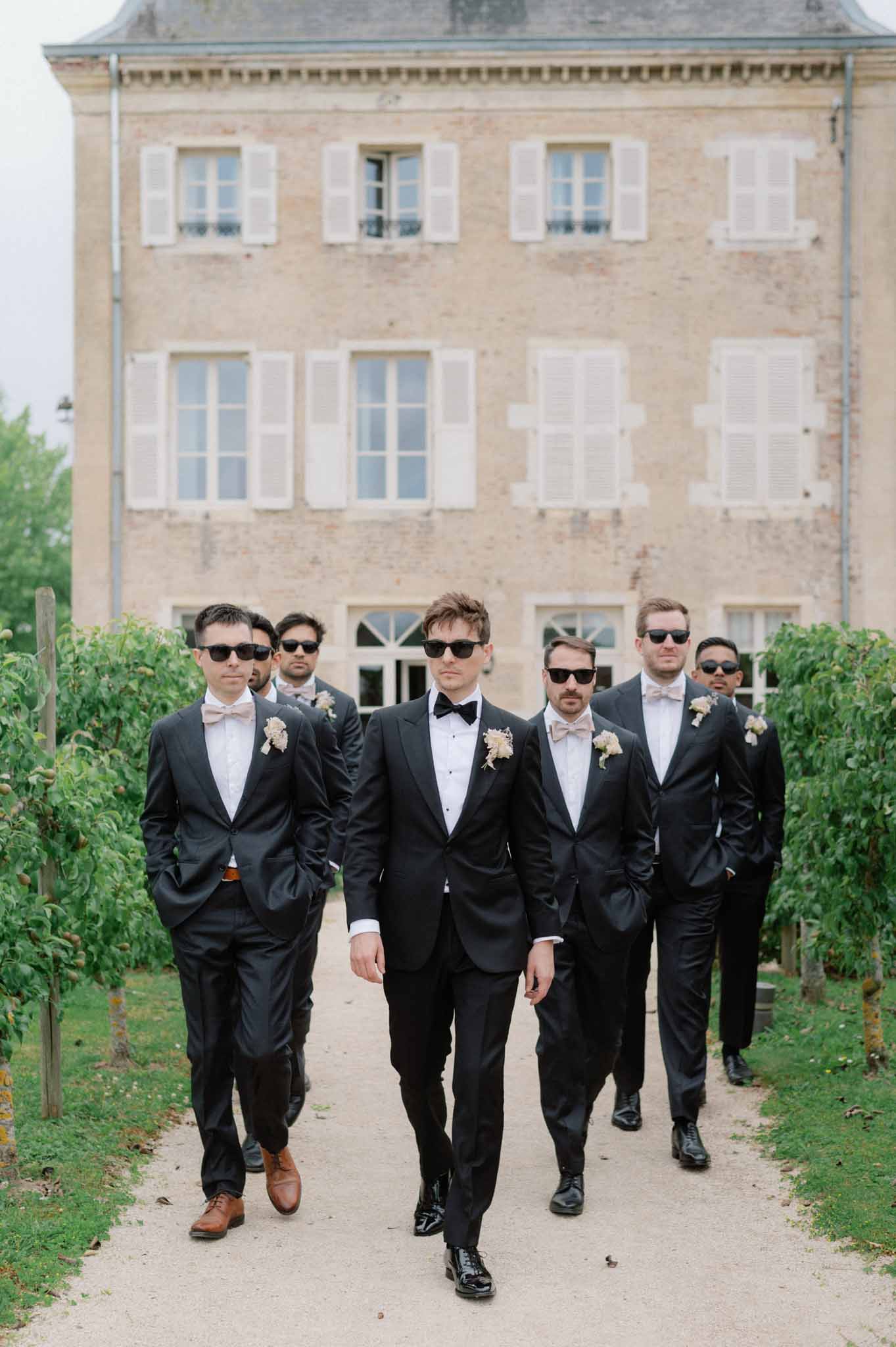 Groom and five groomsmen in black tuxedos walking on gravel path toward camera at a French chateau