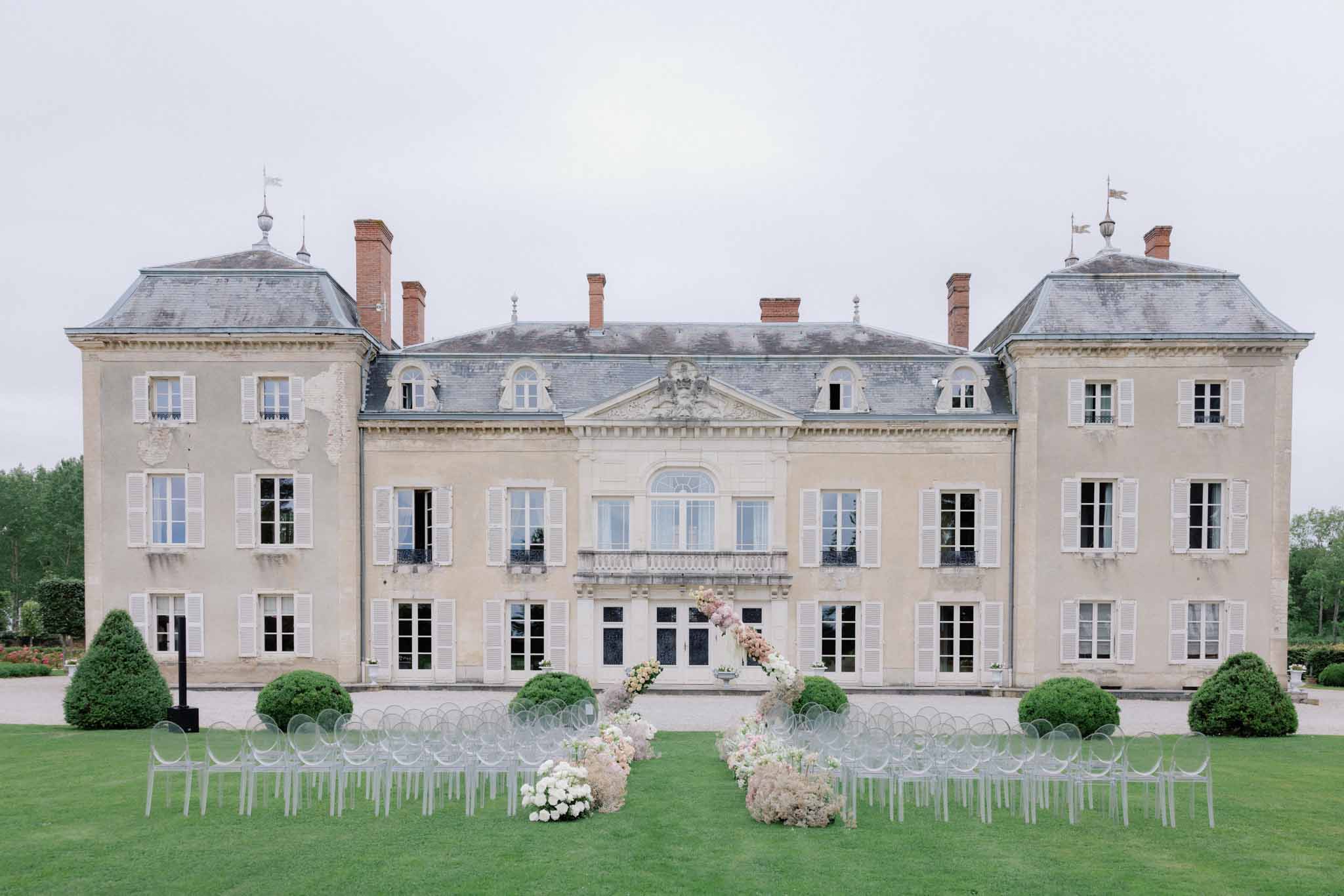 Outdoor ceremony setup on chateau lawn with acrylic chairs, white rose and pampas grass aisle arrangements