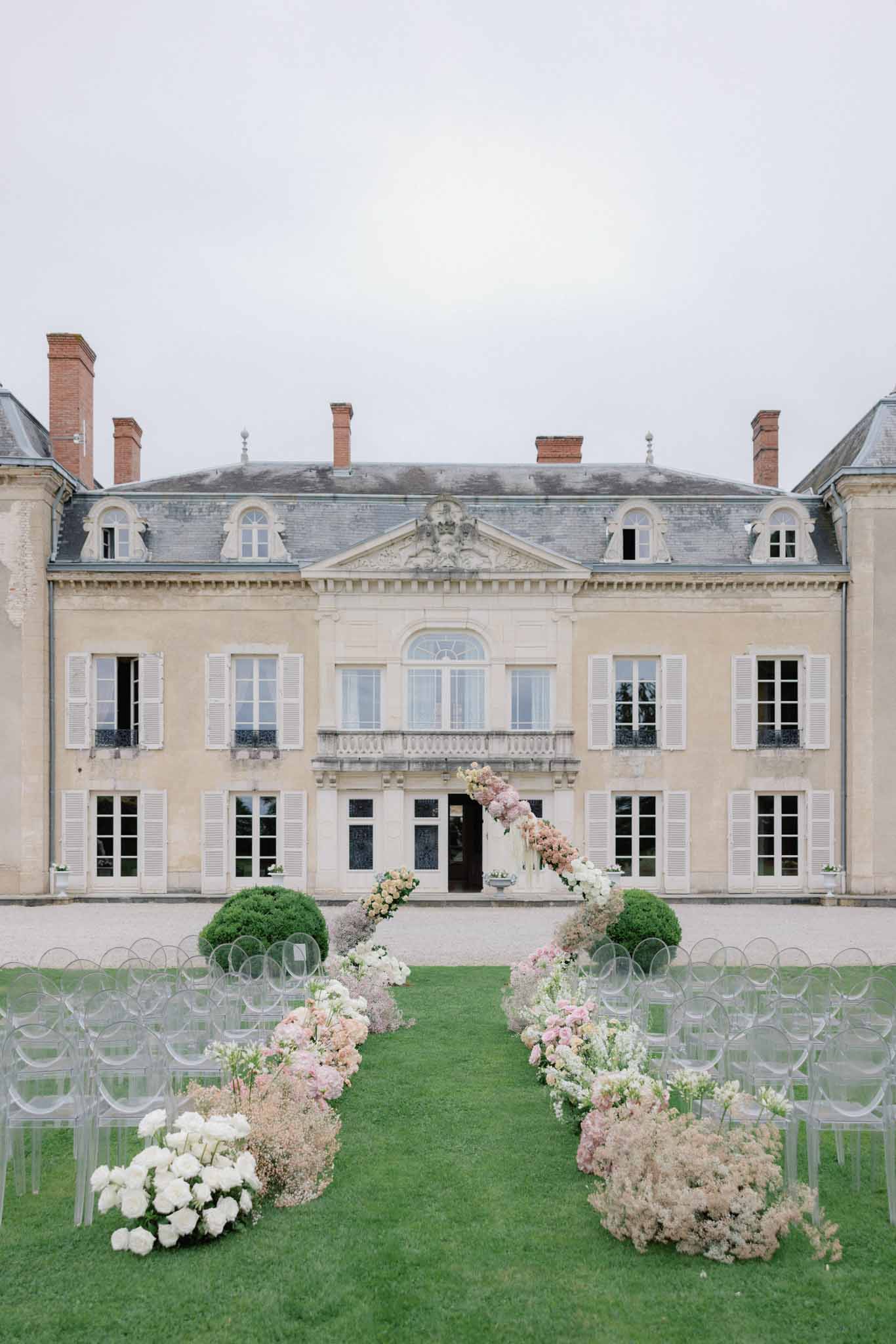 Ceremony aisle lined with blush roses and white hydrangeas in transparent acrylic chairs in courtyard of French chÃ¢teau