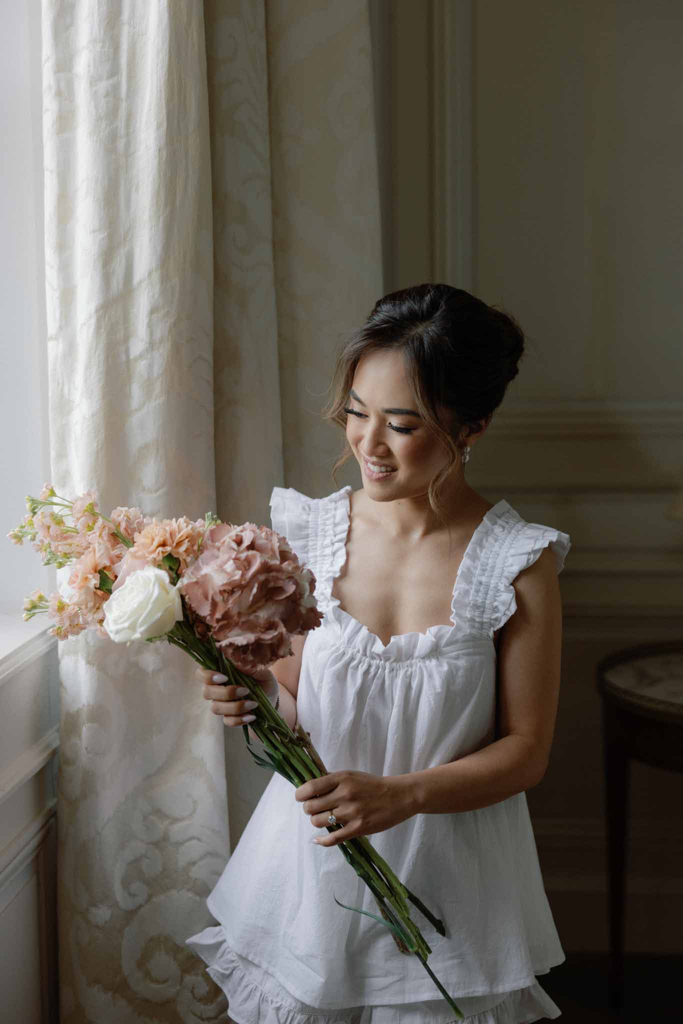 Bride in white flutter-sleeve dress smiling at bouquet of dusty rose carnations and ivory garden roses
