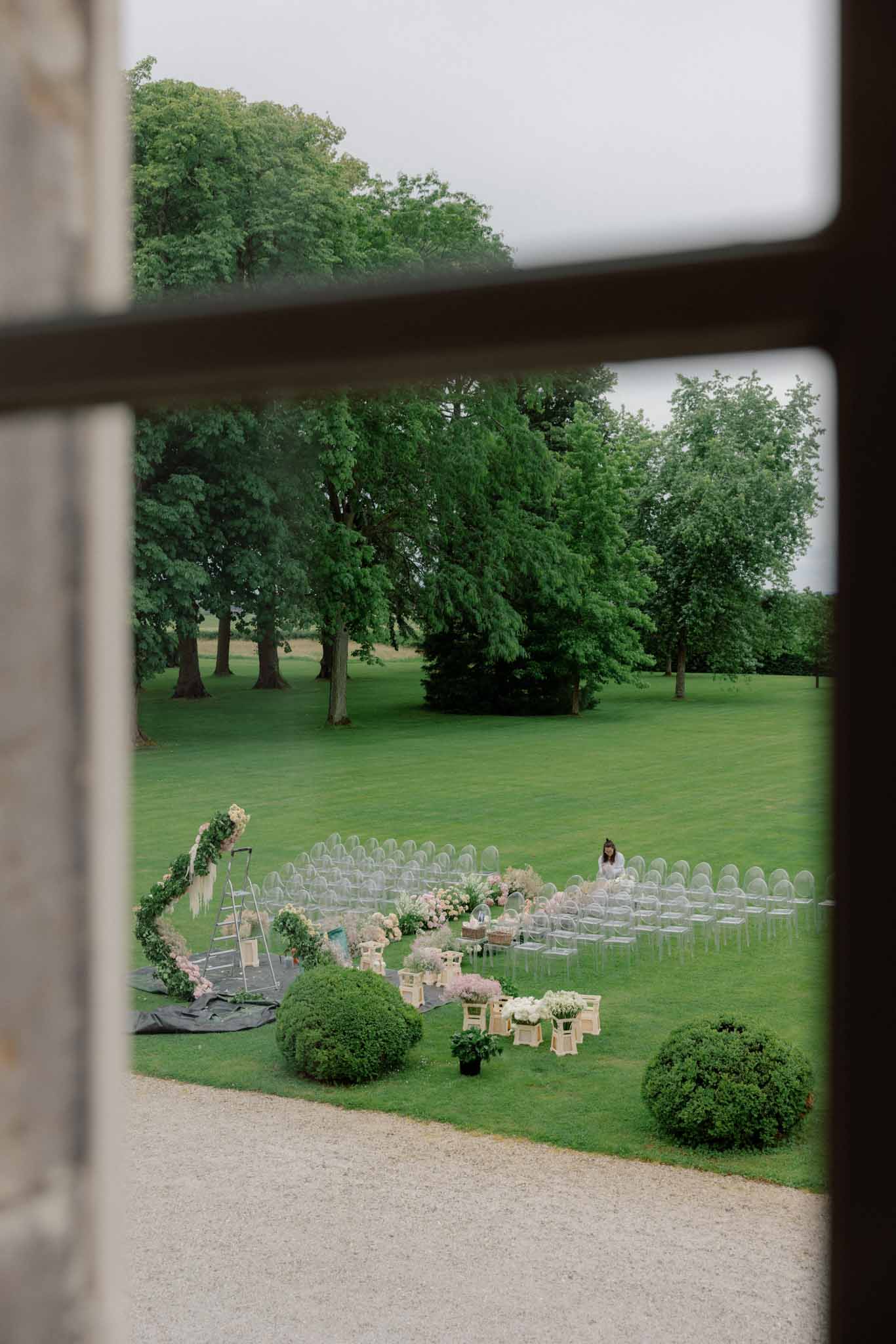 Outdoor ceremony setup on manicured lawn viewed through open window with clear Chiavari chairs