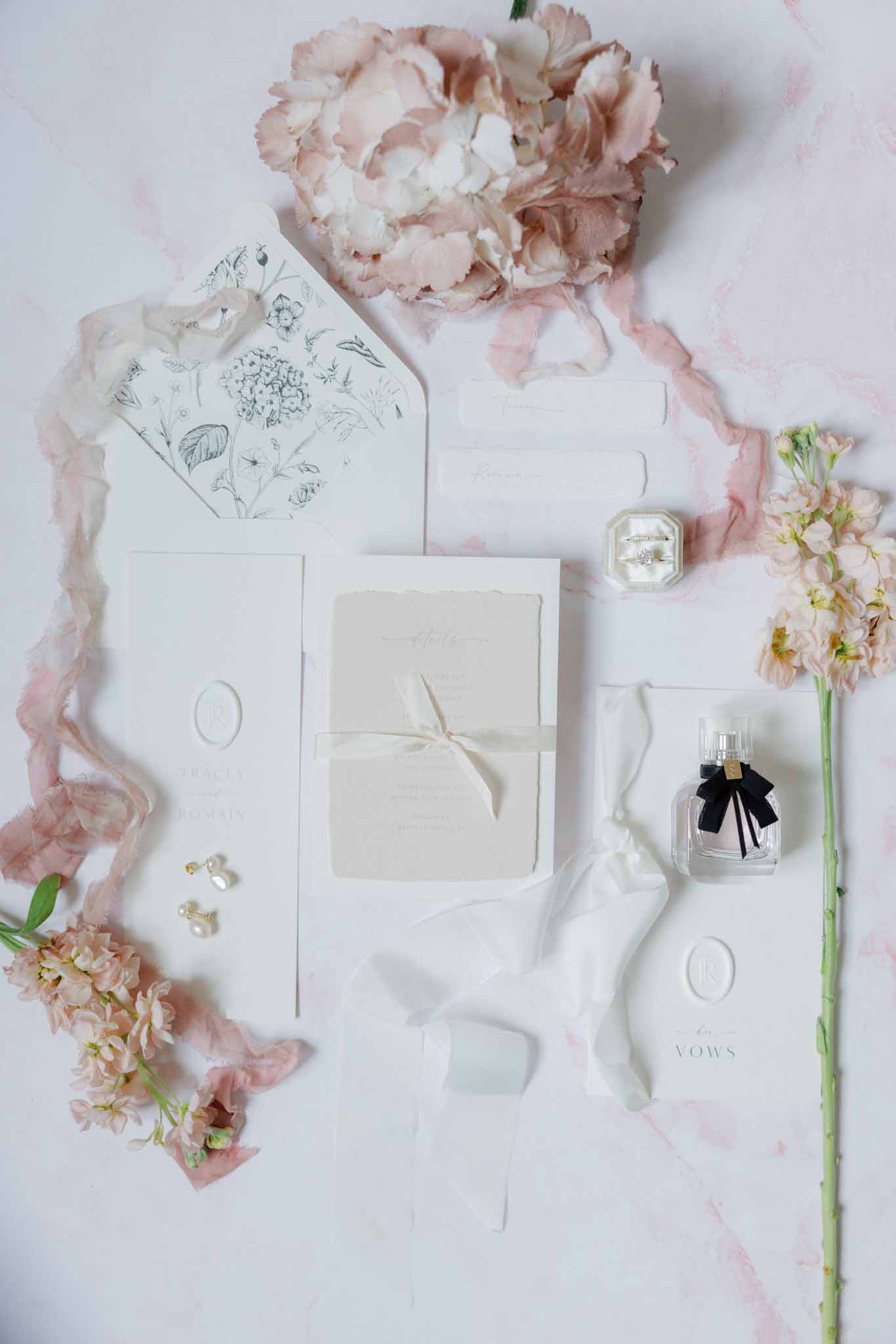 Wedding stationery flat lay with monogrammed invitations, gold earrings, diamond ring, and dusty rose peonies