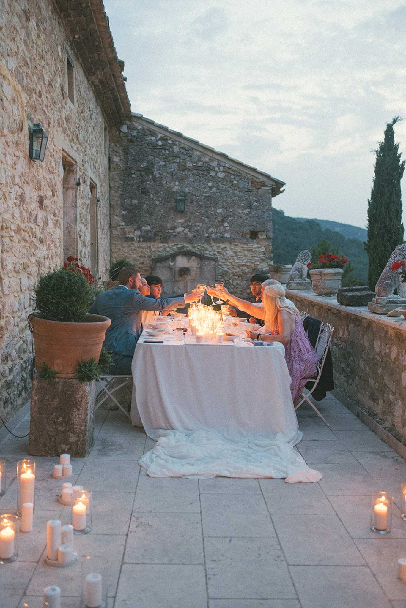 Couple and guests at candlelit round table on stone terrace at dusk raising glasses in toast, cypress trees behind