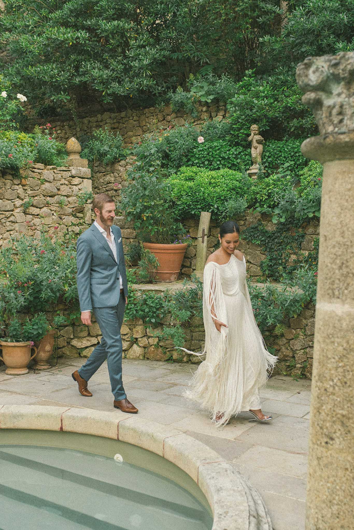 Bride and groom walking through Mediterranean garden courtyard with ivy-covered walls and classical statuary