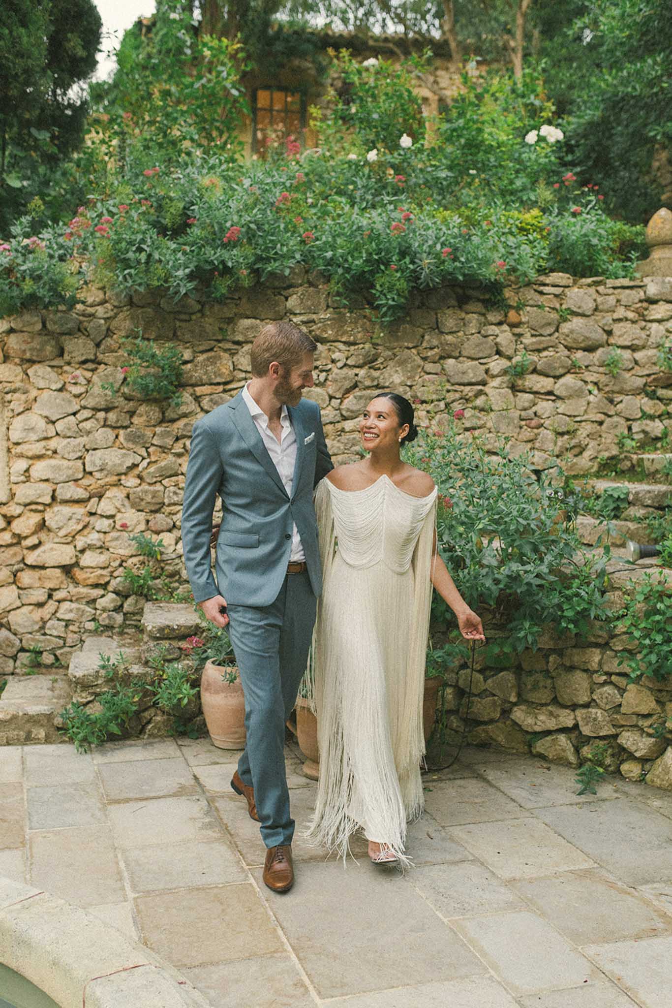 Bride and groom walking together through a Mediterranean garden courtyard with flowering shrubs and stone walls