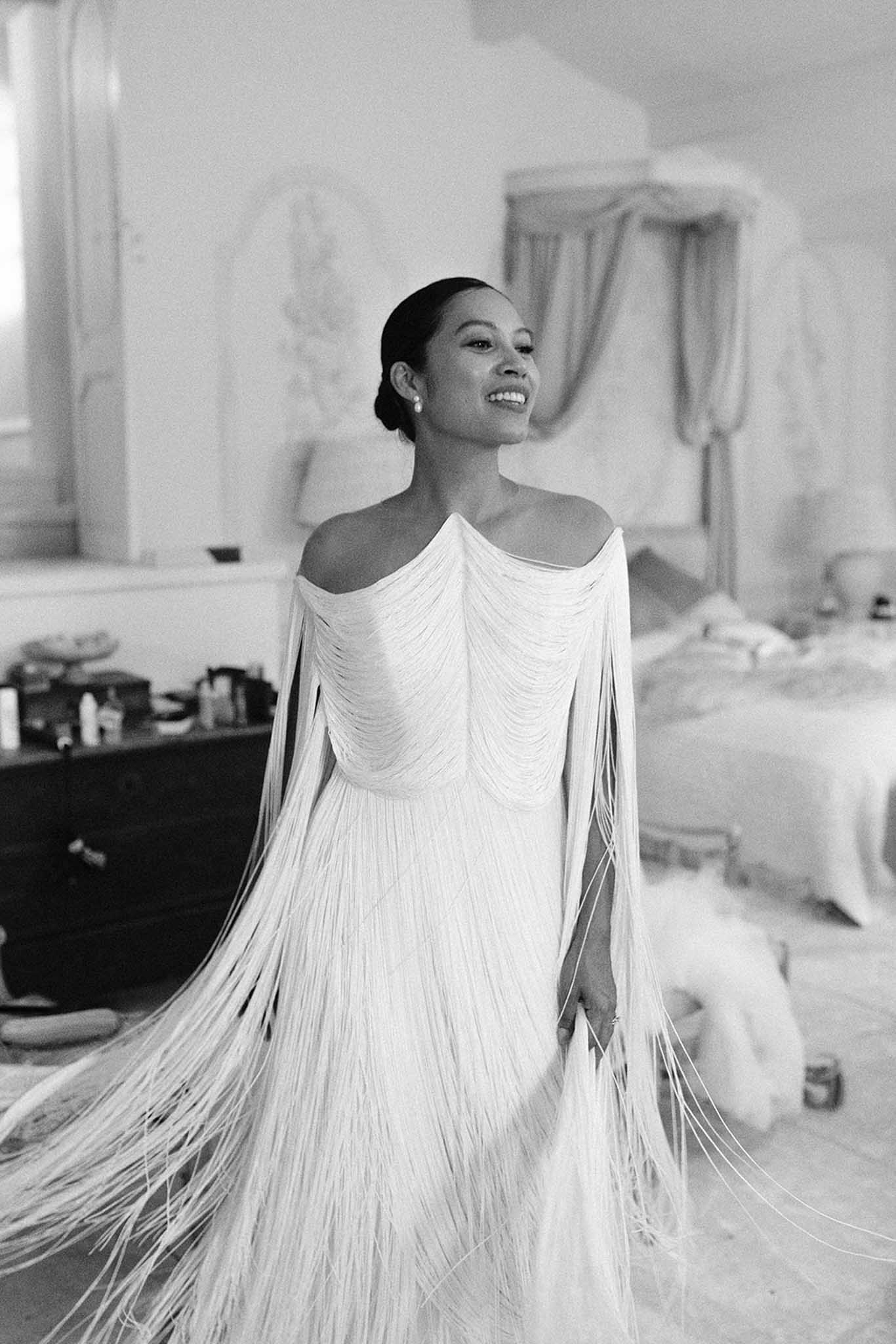 Black and white portrait of smiling bride in draped off-shoulder gown with long fringed cape in bridal suite