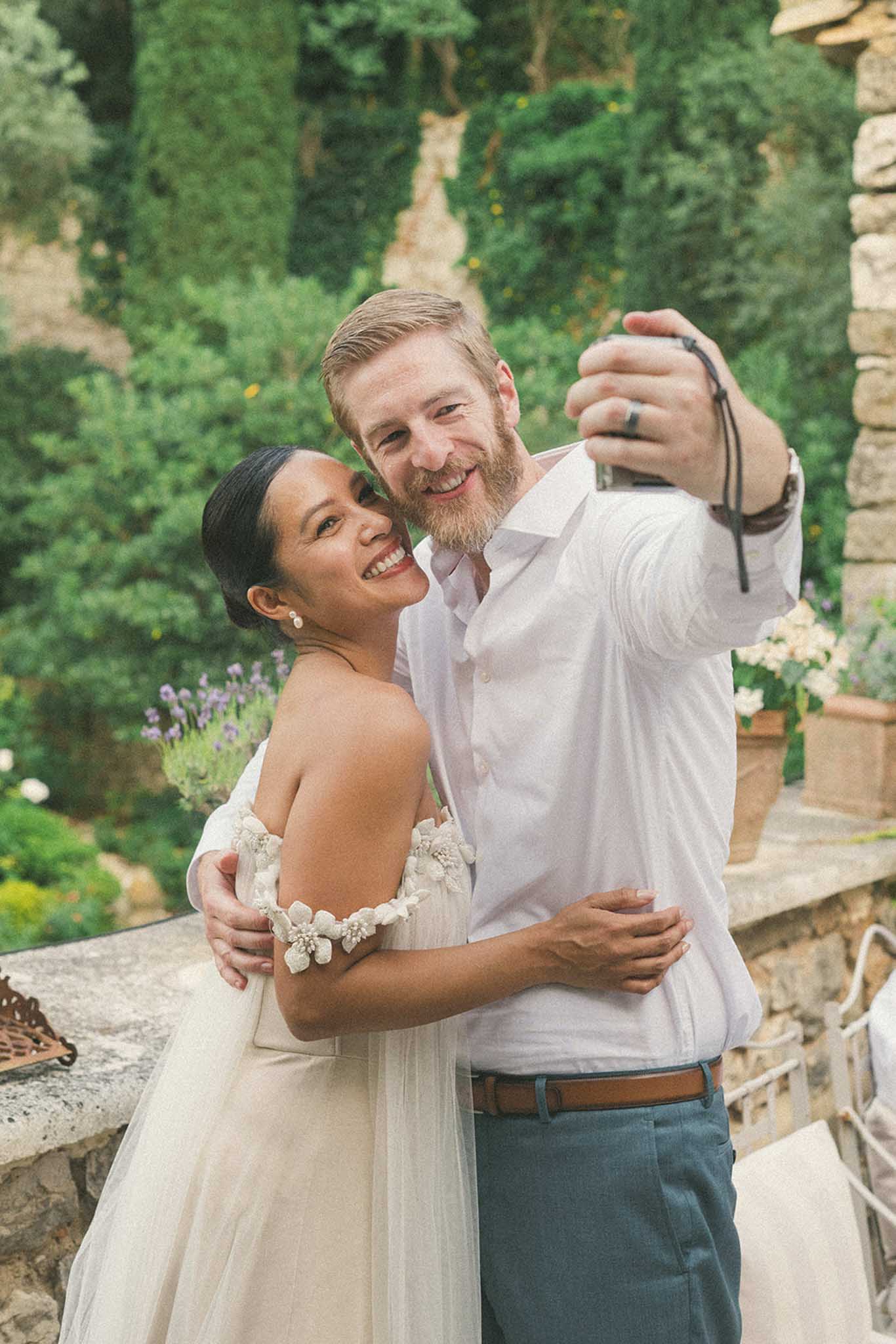 Bride in off-shoulder ivory tulle gown and groom in gray linen shirt taking a selfie in a garden with lavender and ivy-covered stone walls.