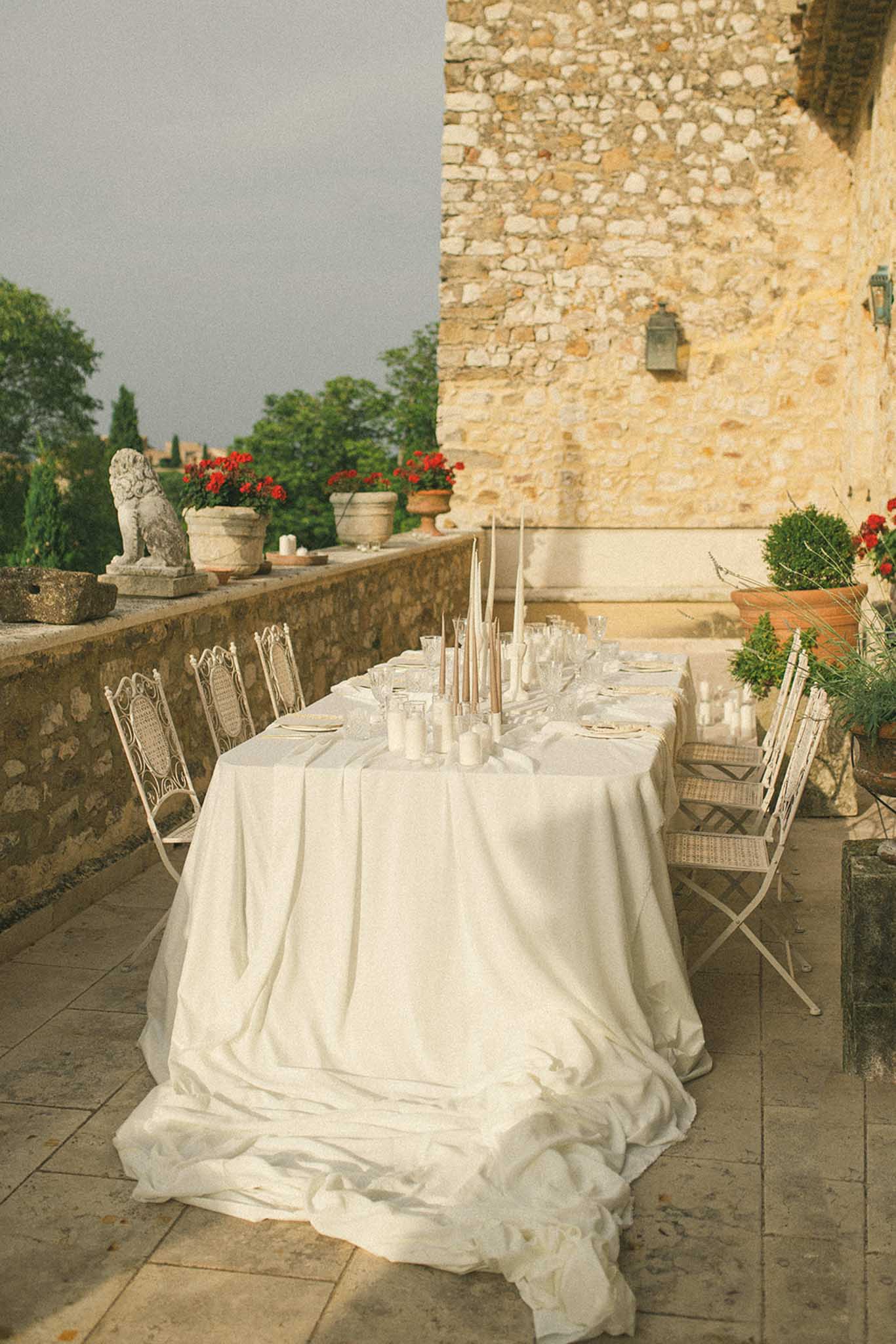 Reception table on stone terrace of historic chÃ¢teau with rattan chairs, red geraniums and lion statue in background
