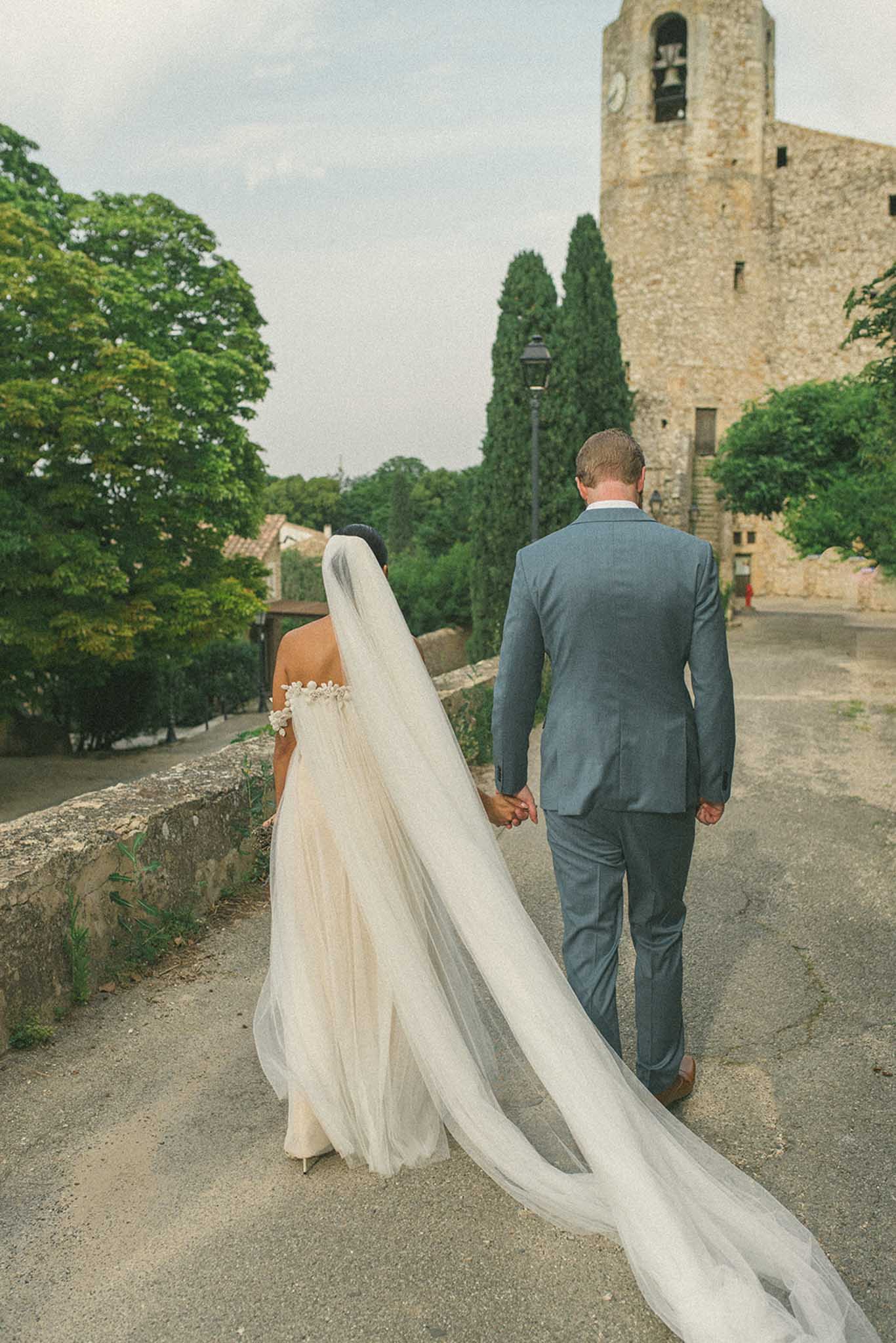 Bride and groom walking from medieval stone church with trailing veil and cypress trees