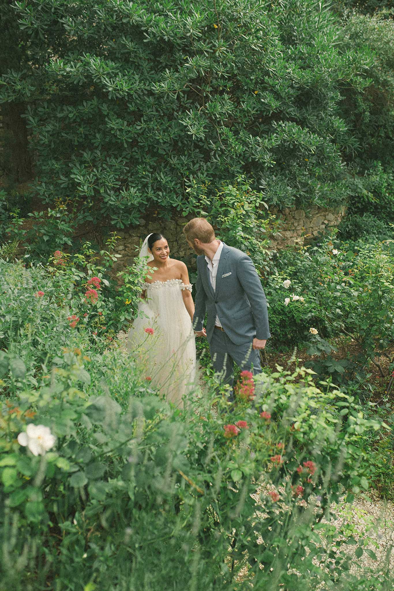Bride in ivory tulle gown and groom in grey suit walking through garden pathway with climbing ivy and red blooms