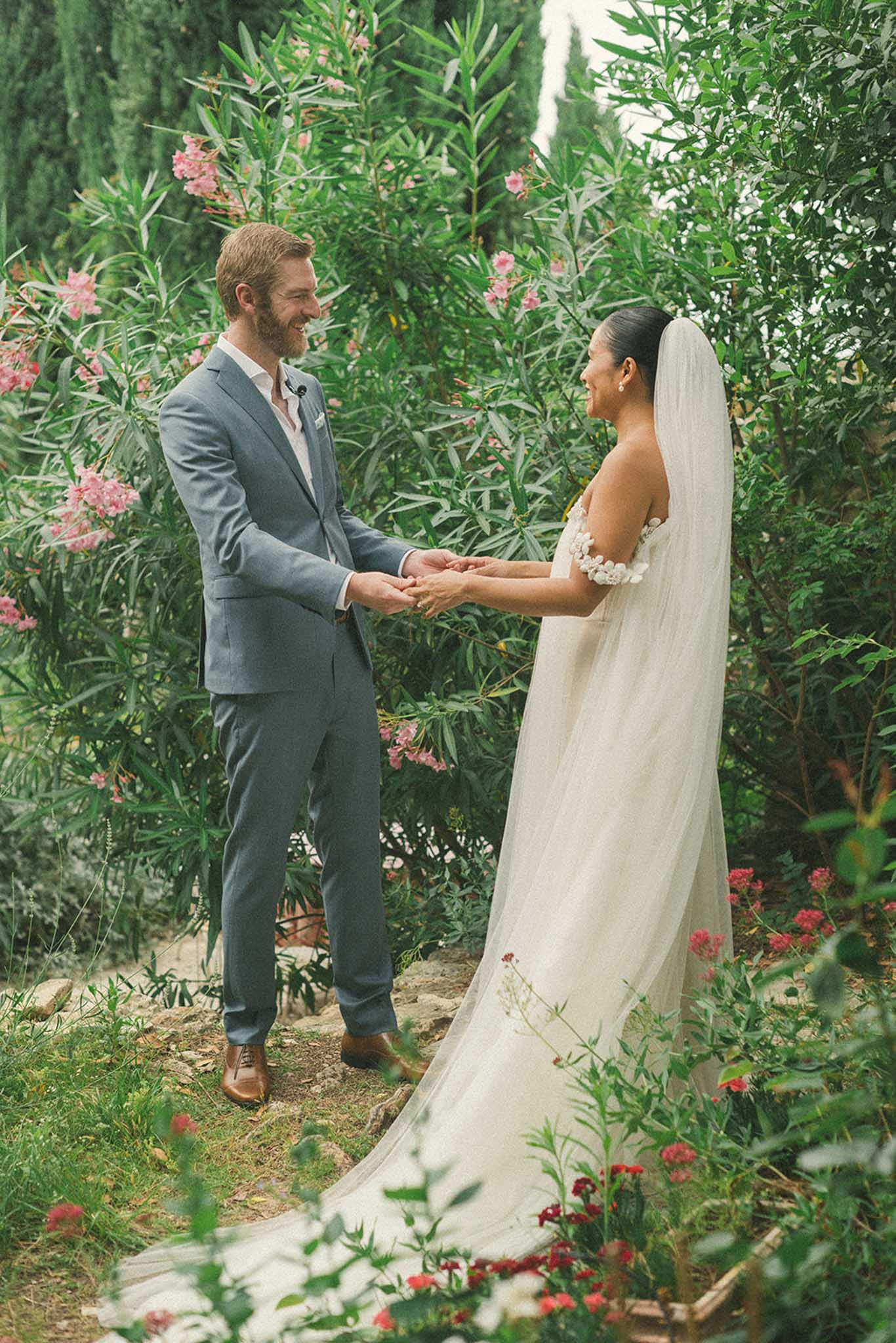 Groom in blue suit and bride exchanging vows holding hands in a garden with cypress trees and pink flowers