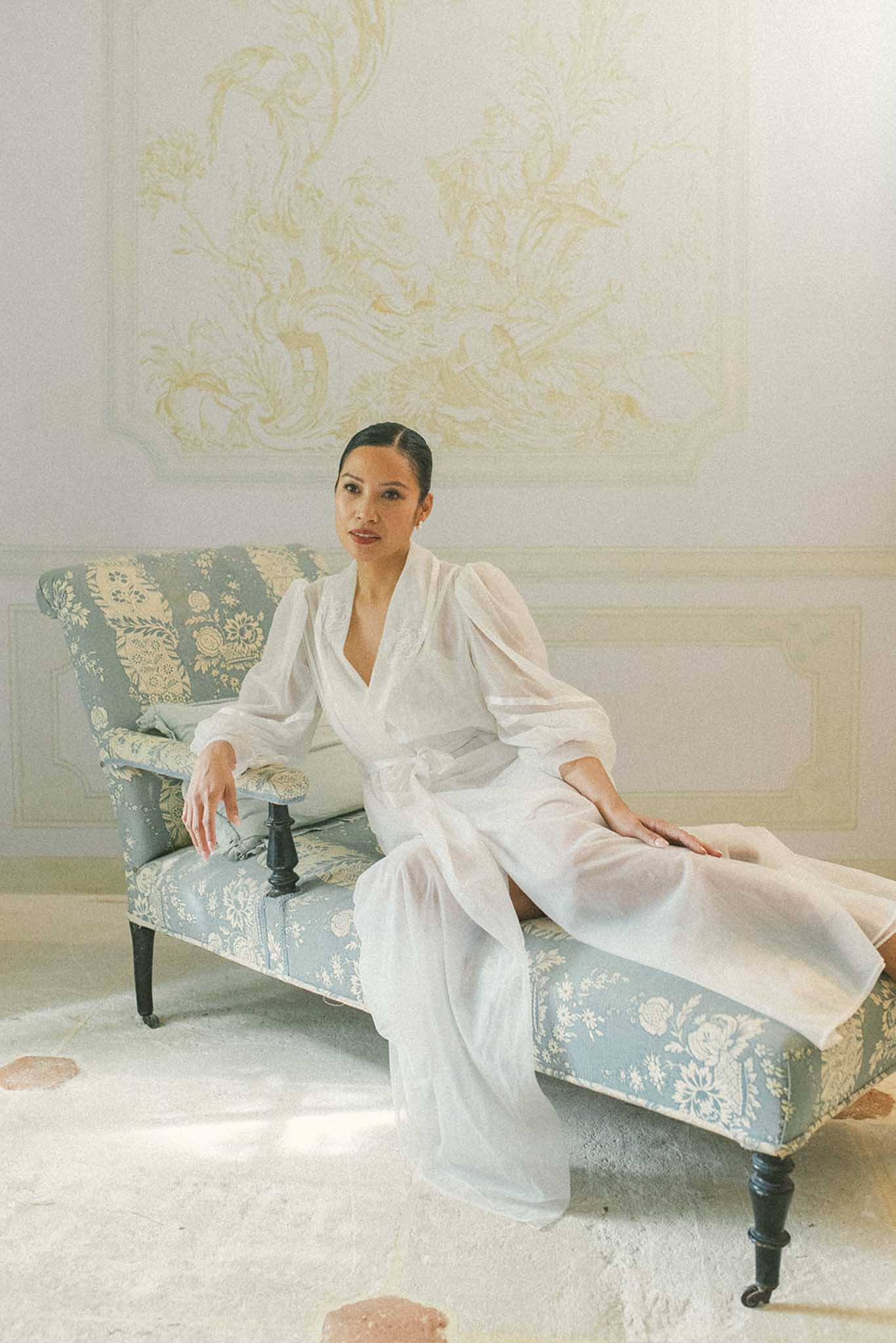 Bride in sheer organza gown with puffed sleeves seated on antique settee in gold-panelled chateau interior