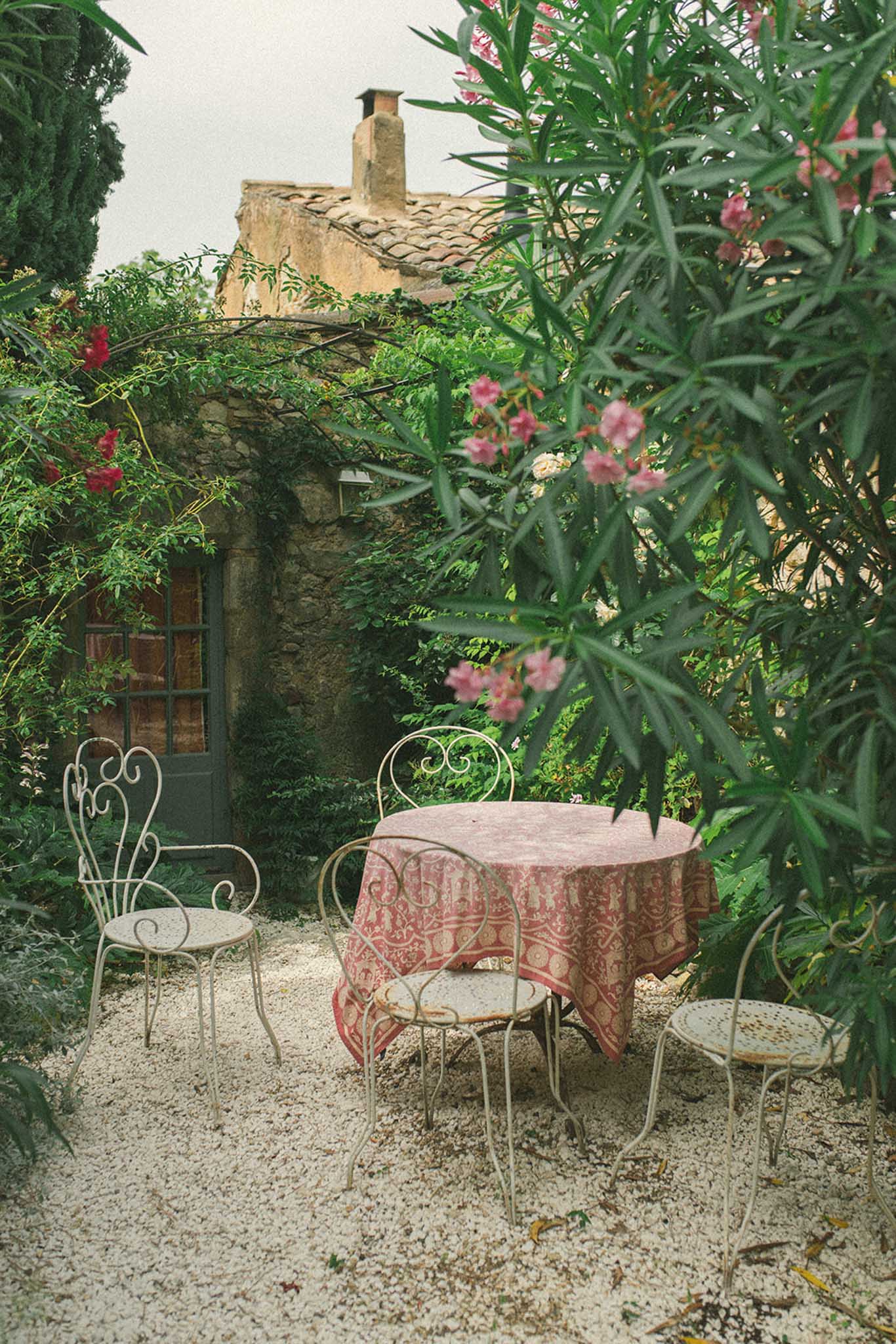 Stone building with terracotta roof framed by pink climbing vines with bistro table and iron chairs in courtyard