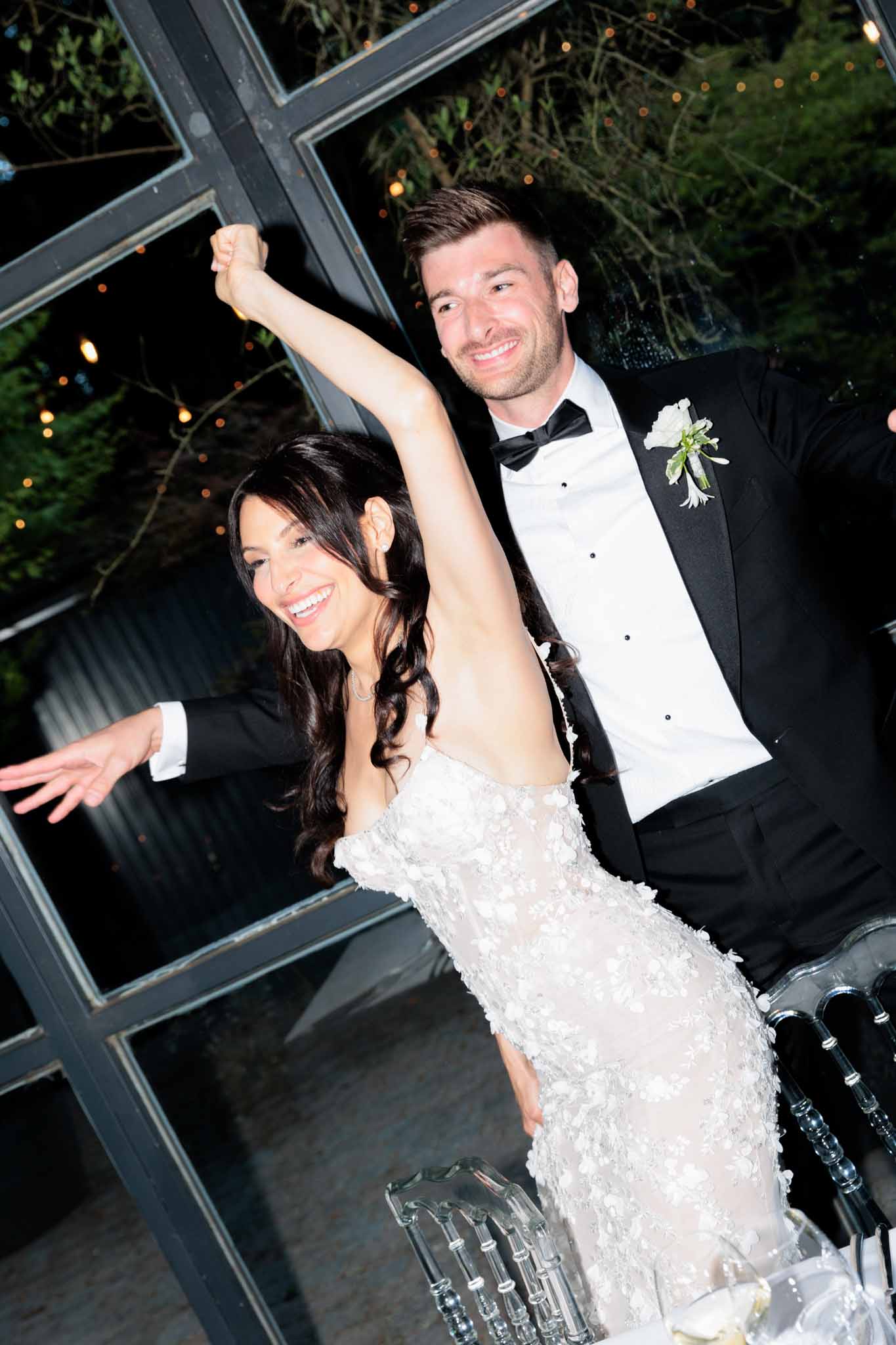 The bride and groom are captured mid-celebration, likely during their reception entrance or first dance, both laughing and raising their joined hands in the air. The setting appears to be an indoor venue with large dark-framed glass windows or doors, with warm bistro string lights and foliage visible through the glass in the background. The bride wears a form-fitting, off-the-shoulder white gown covered in three-dimensional floral appliqués, with her dark hair in loose waves; the groom wears a classic black tuxedo with a black bow tie and a white floral boutonnière. The shot is a close-up portrait at a slight angle, with acrylic chairs partially visible in the lower foreground, suggesting a modern classic decor style.