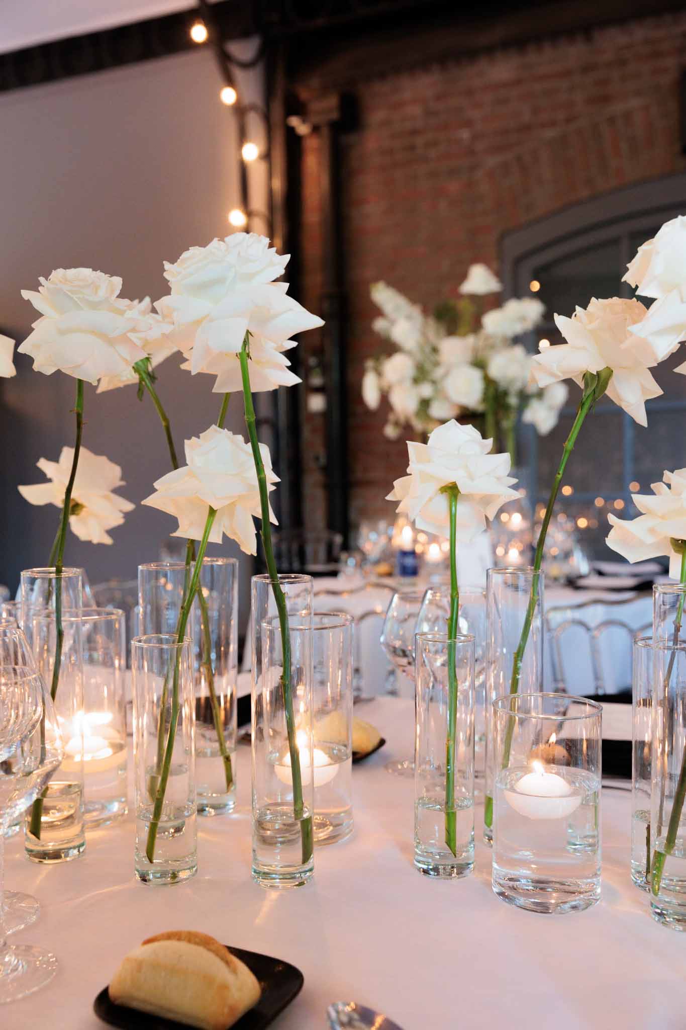 Close-up detail shot of a wedding reception tablescape inside an industrial-style venue with exposed brick walls and Edison bulb string lights. The centerpiece consists of multiple individual white roses on long green stems, each placed in separate tall clear glass cylinder vases, mixed with small floating candle holders containing lit white tea lights. The table is covered in a white linen cloth and set with wine glasses and black bread plates holding sliced baguette. In the background, additional round guest tables are visible, similarly decorated with white floral arrangements and candlelight, with ghost-style chairs partially visible. The overall decor palette is white and clear glass with a modern, minimal aesthetic.