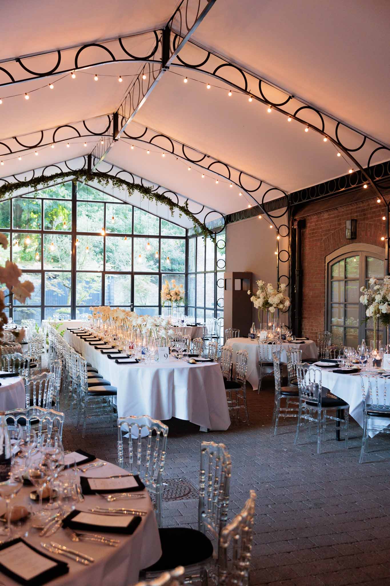 Orangery reception with ghost chairs, fairy lights, tall white floral centerpieces, and iron windows
