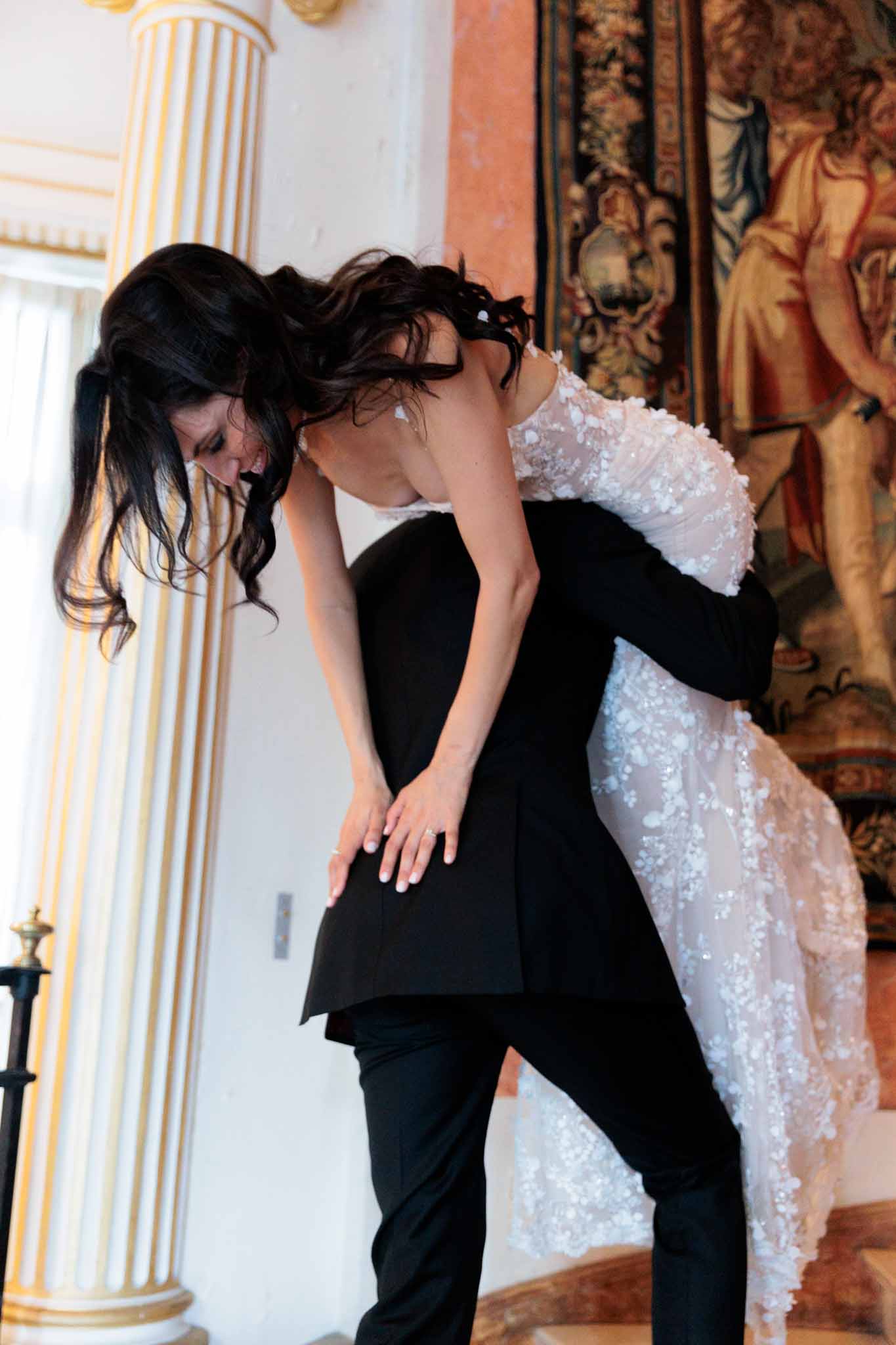 The groom, dressed in a black tuxedo, lifts the bride over his shoulder in a playful moment inside a formal interior space. The bride wears a white floral appliqué lace gown with a sheer bodice and a flowing skirt, her dark hair loosely falling around her face as she laughs. The setting features gold-trimmed classical columns, a marble floor, and a large ornate tapestry with figurative imagery on the wall behind them. The shot is a mid-range portrait capturing the couple's movement and energy in a classic French château-style interior.