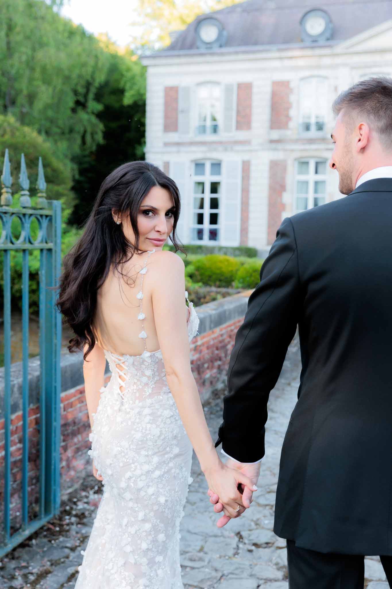 A couple portrait taken outdoors on a cobblestone path beside ornate blue iron gates, with a French château featuring brick and stone facade visible in the background. The bride, who has long dark hair worn down, glances back over her shoulder toward the camera while holding the groom's hand; she wears a form-fitting white gown with all-over 3D floral appliqué, a deep open back with corset lacing, and a delicate crystal or pearl back necklace draping down the spine. The groom, partially turned away from the camera, wears a classic black tuxedo jacket. The composition is a mid-range portrait shot with the couple in the foreground and the château softly out of focus behind them.
