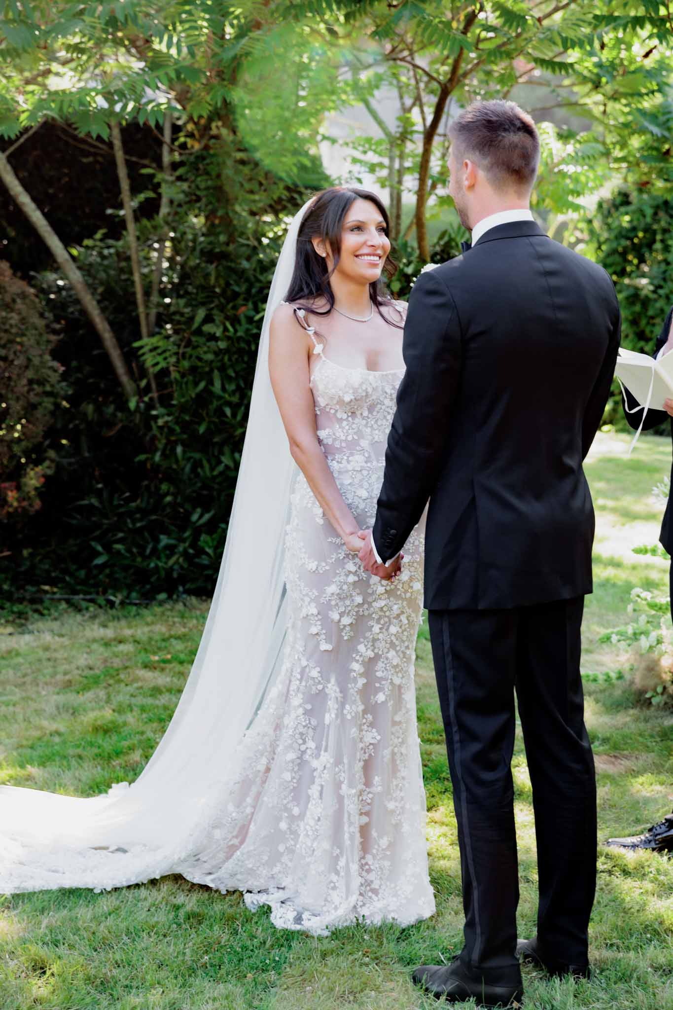 Bride in sheer floral applique gown and cathedral veil holding hands with groom during outdoor ceremony vows