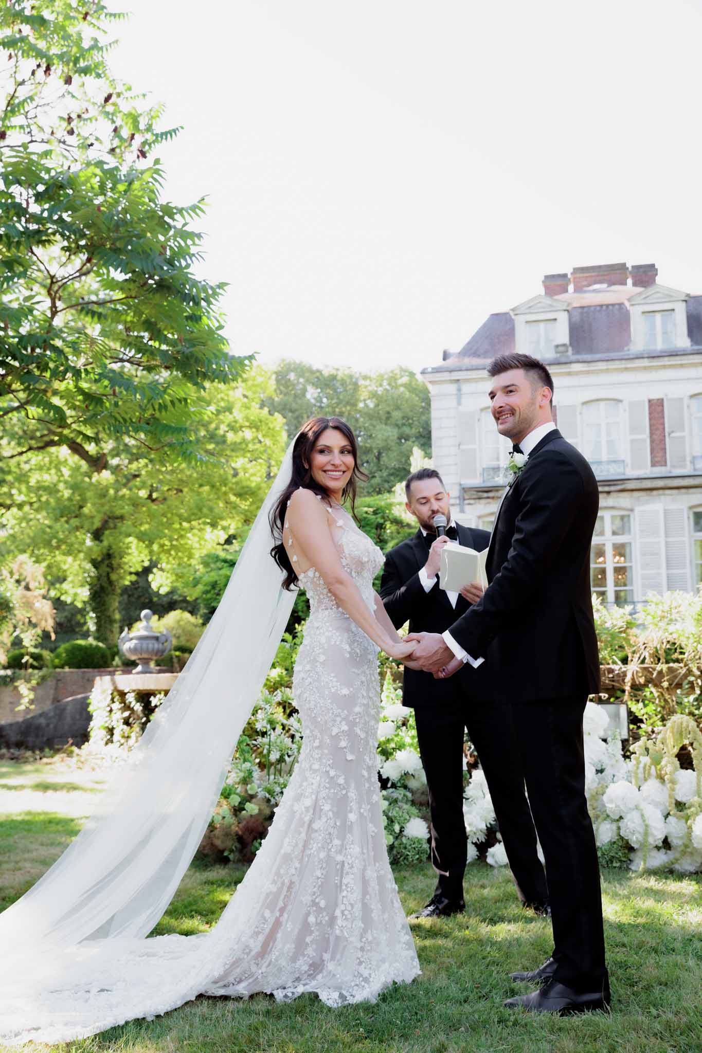 Bride in lace gown and groom at ceremony with white hydrangea backdrop before chateau facade