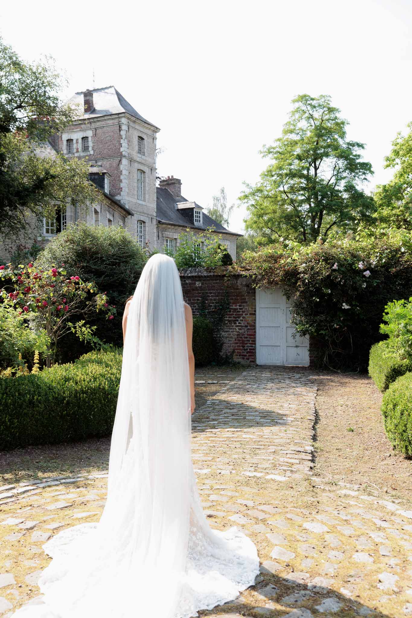 Bride seen from behind wearing lace-edged cathedral train and veil in walled garden of French brick manor house