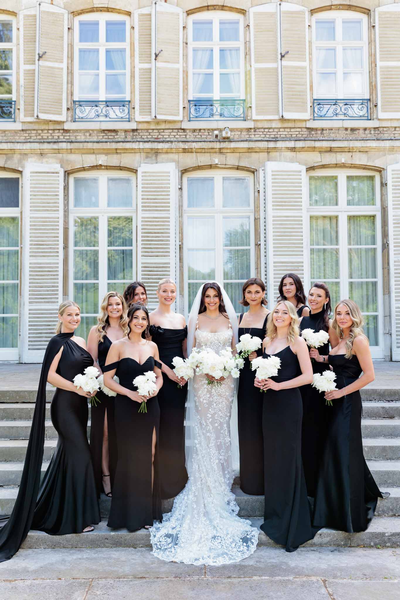 A bridal party portrait taken outdoors on the stone steps of a French château, with the building's cream limestone facade, arched windows, and white shuttered doors filling the background. The bride stands at center wearing a fitted, floor-length lace gown with intricate floral appliqué and a long lace cathedral train, paired with a white veil, and holds a large bouquet of white peonies. She is flanked by ten bridesmaids dressed in mismatched black floor-length gowns in various styles — including off-the-shoulder, cape, halter neck, spaghetti strap, and square-neck silhouettes — each holding a smaller bouquet of white peonies. The overall color palette is a high-contrast black and white, creating a modern and polished aesthetic. This is a wide group portrait shot with all subjects facing the camera.