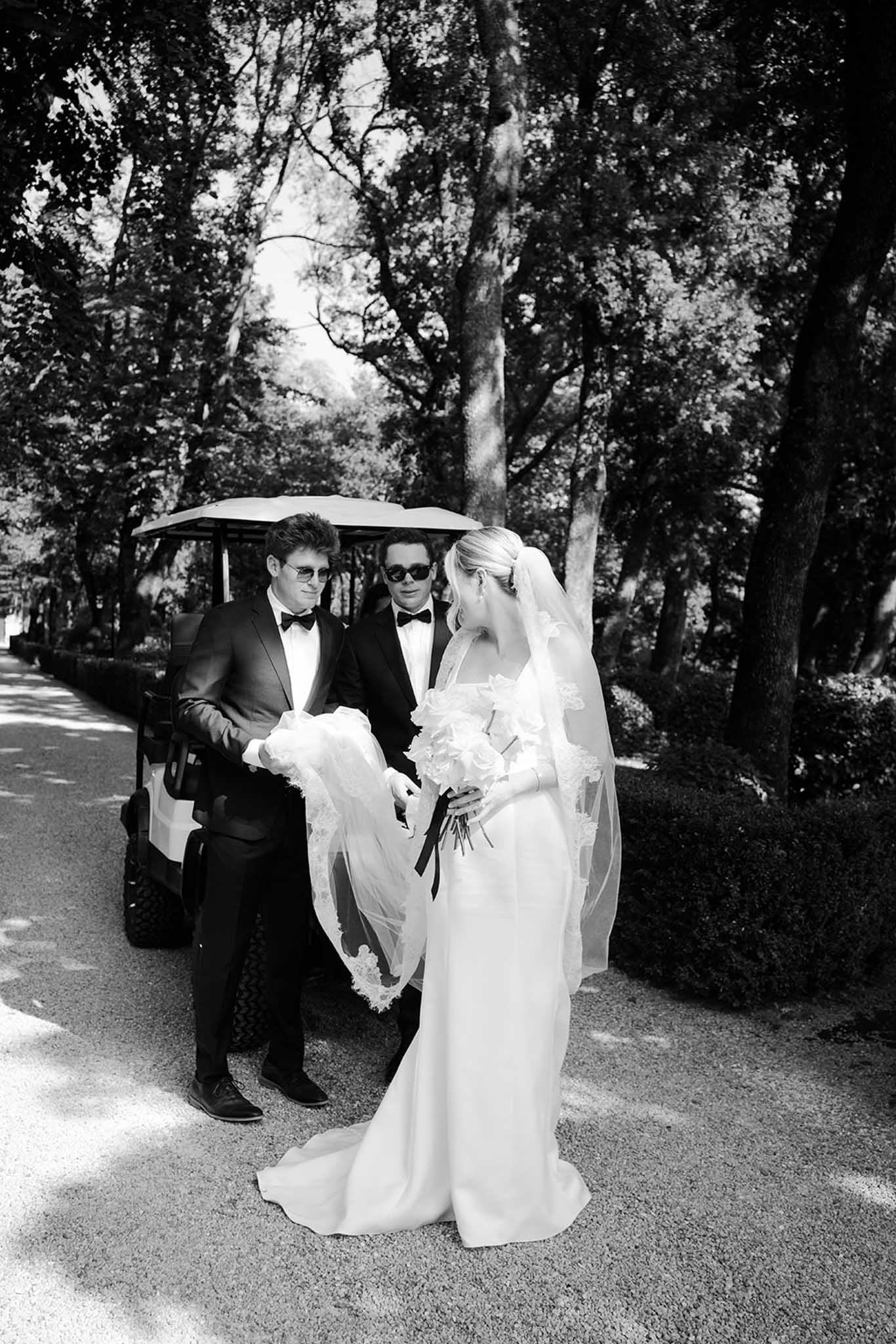 Bride in column gown with veil flanked by two groomsmen in black tuxedos and sunglasses on tree-lined driveway