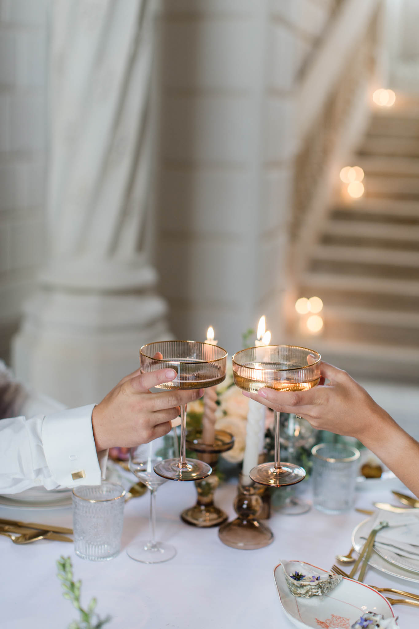 Couple toasting champagne at reception table with botanical plates, gold flatware, and candlelit staircase backdrop