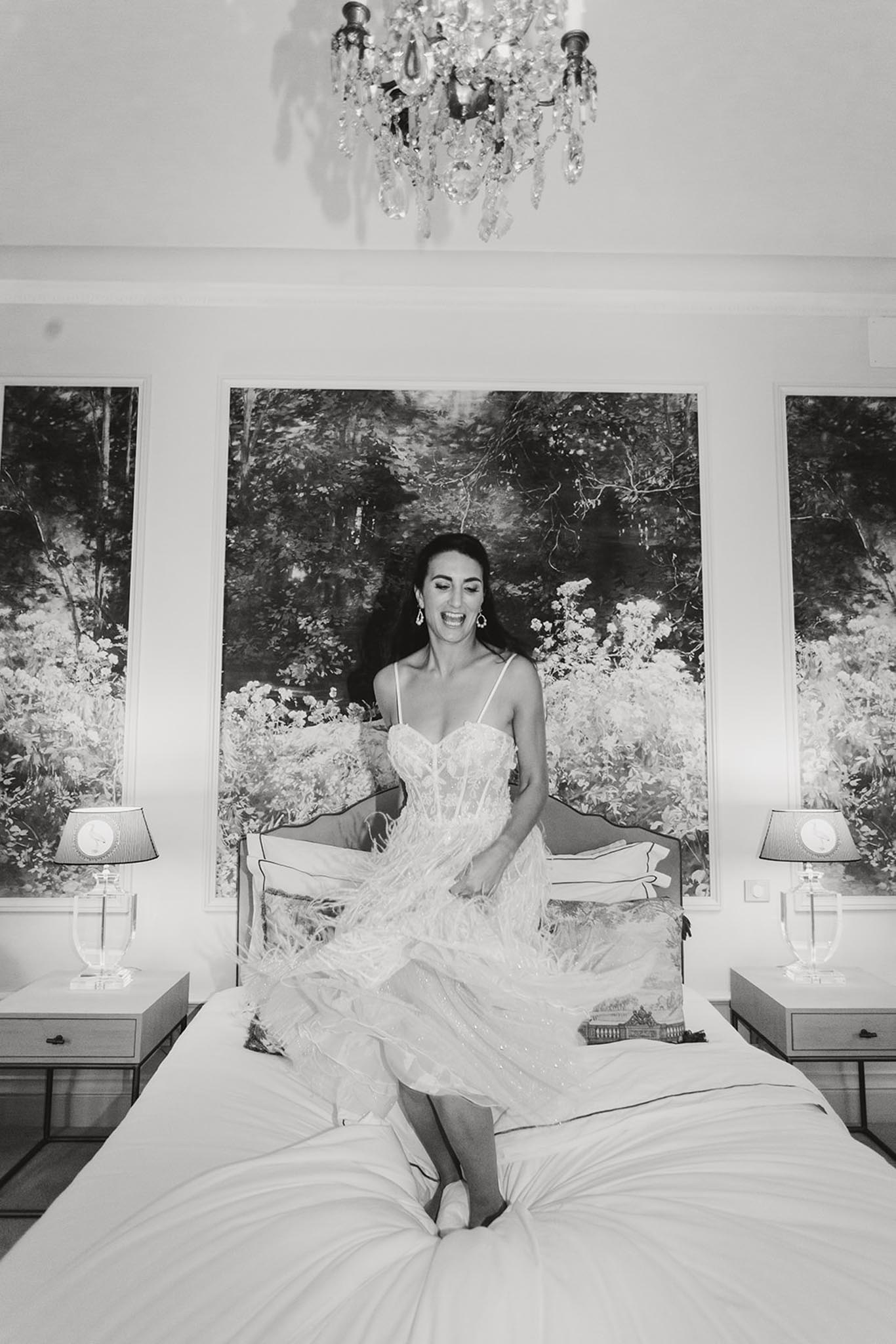 Bride in layered tulle wedding dress seated on white-linen bed in symmetrical bedroom with chandelier, black and white photo