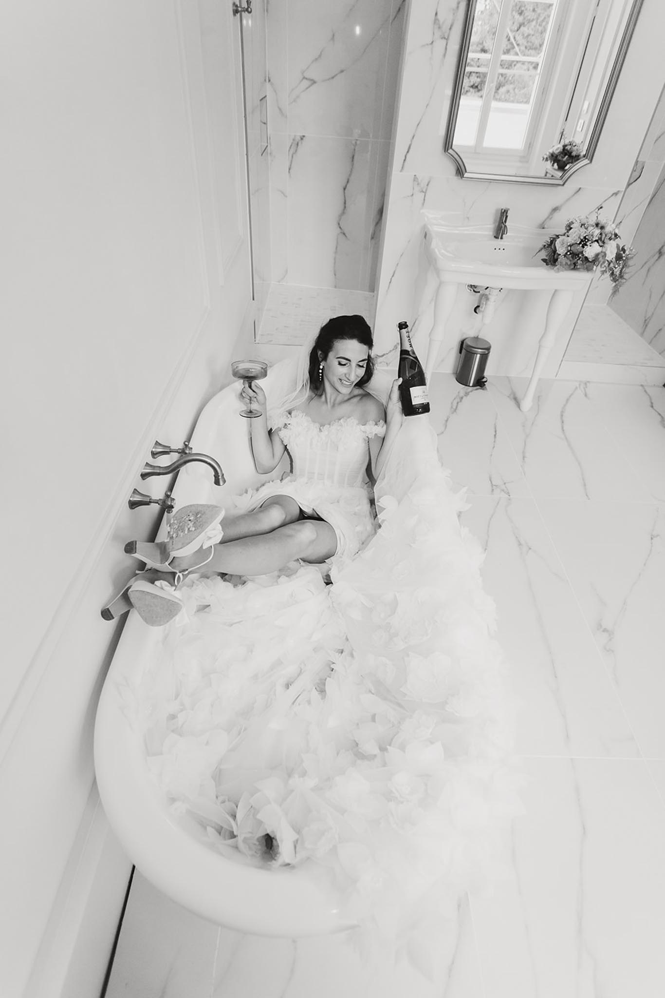 Black-and-white overhead shot of bride in bathtub with champagne bottle wearing off-shoulder white wedding dress