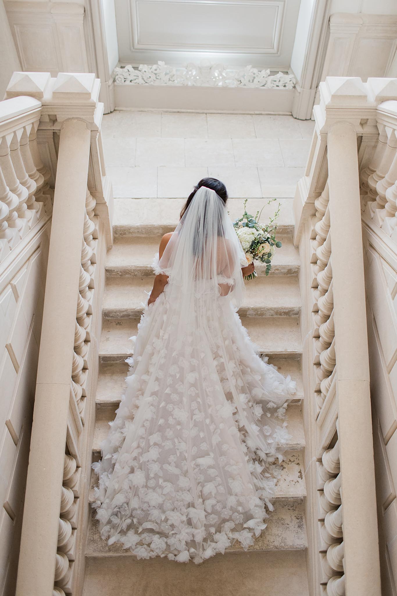 Bride in white ball gown and long veil on a grand stone staircase, viewed from above, neoclassical columns and balustrades visible.