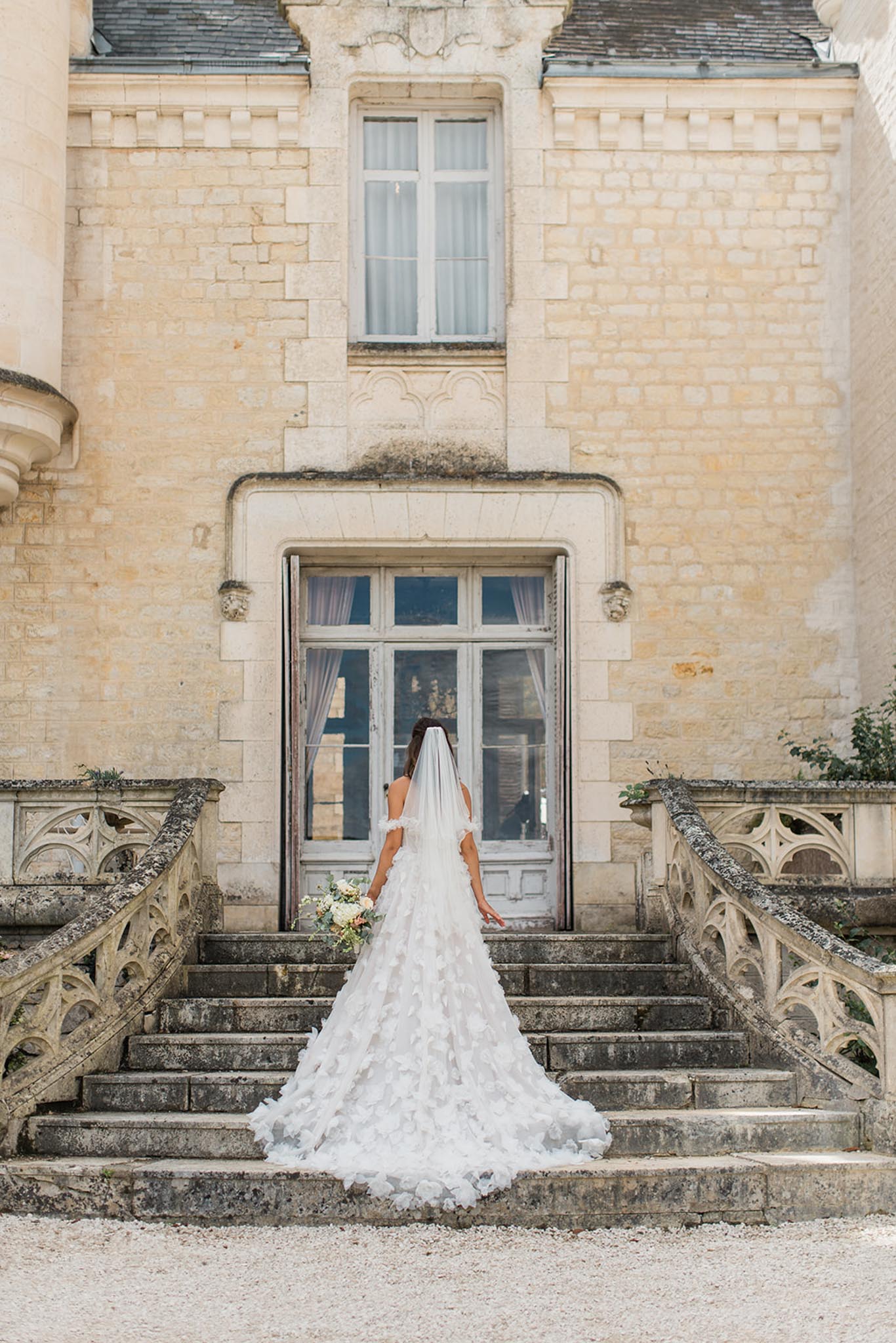 Bride from behind in a white layered gown with cathedral veil trailing down the stone steps of a classical French chÃ¢teau entrance.