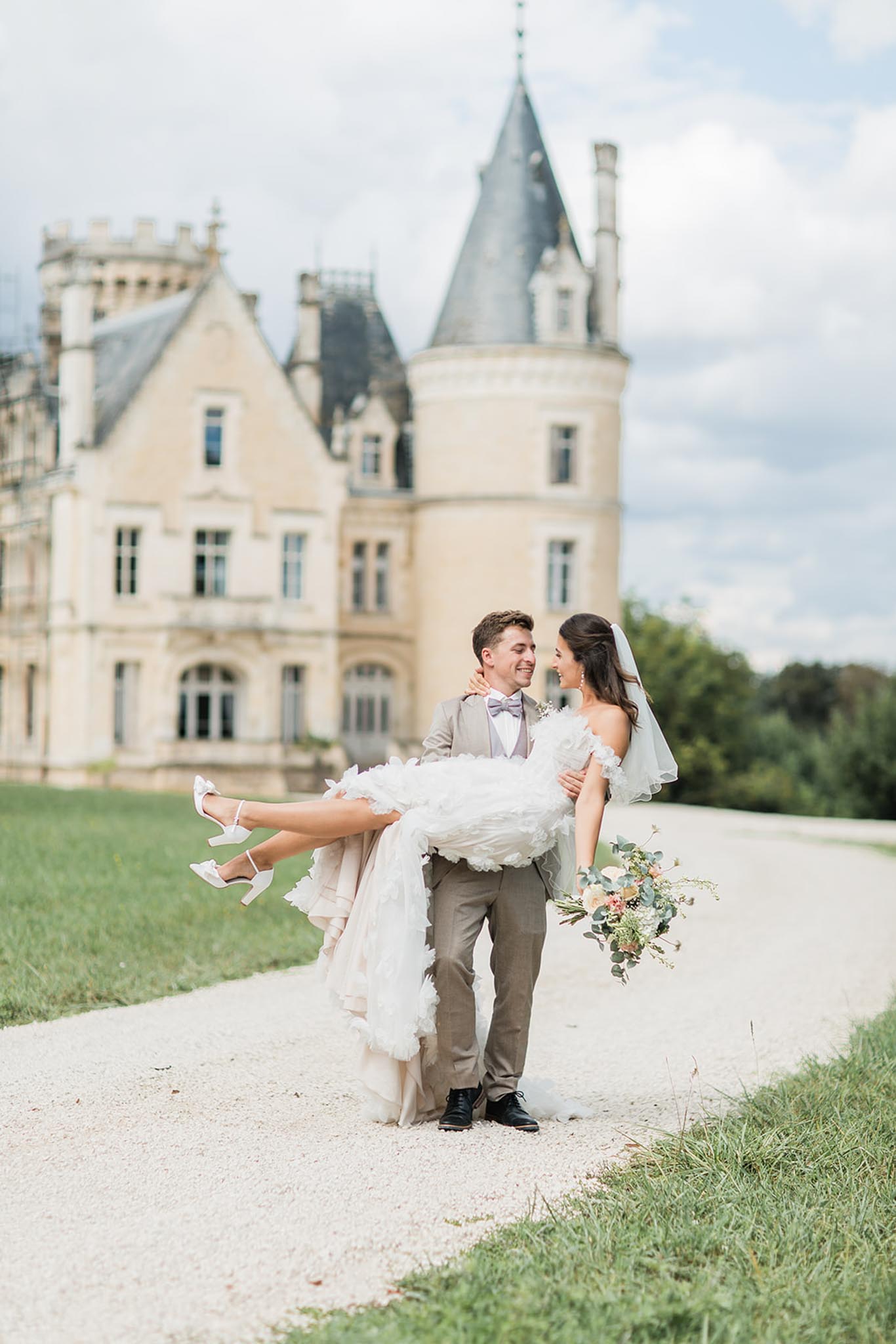 Groom carrying bride on chÃ¢teau grounds, bride in white ruffled gown with chÃ¢teau turret visible behind them