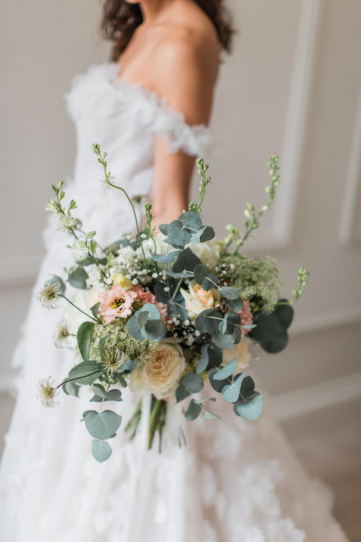 Bride holding garden-style bouquet with cream roses, blush anemones, and eucalyptus against neutral wall