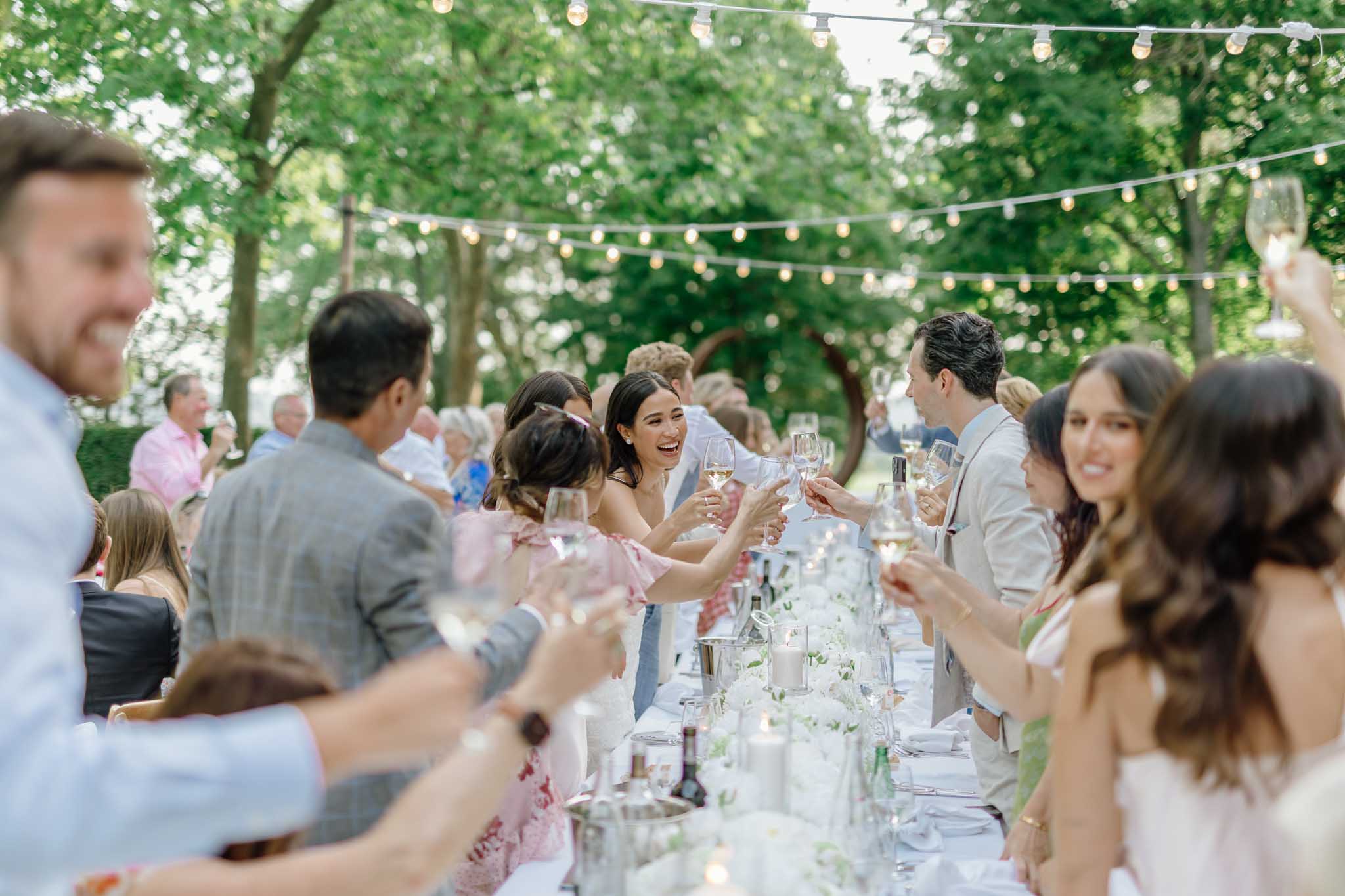 About thirty guests raising glasses in a toast at a long outdoor banquet table under string lights in a tree canopy.