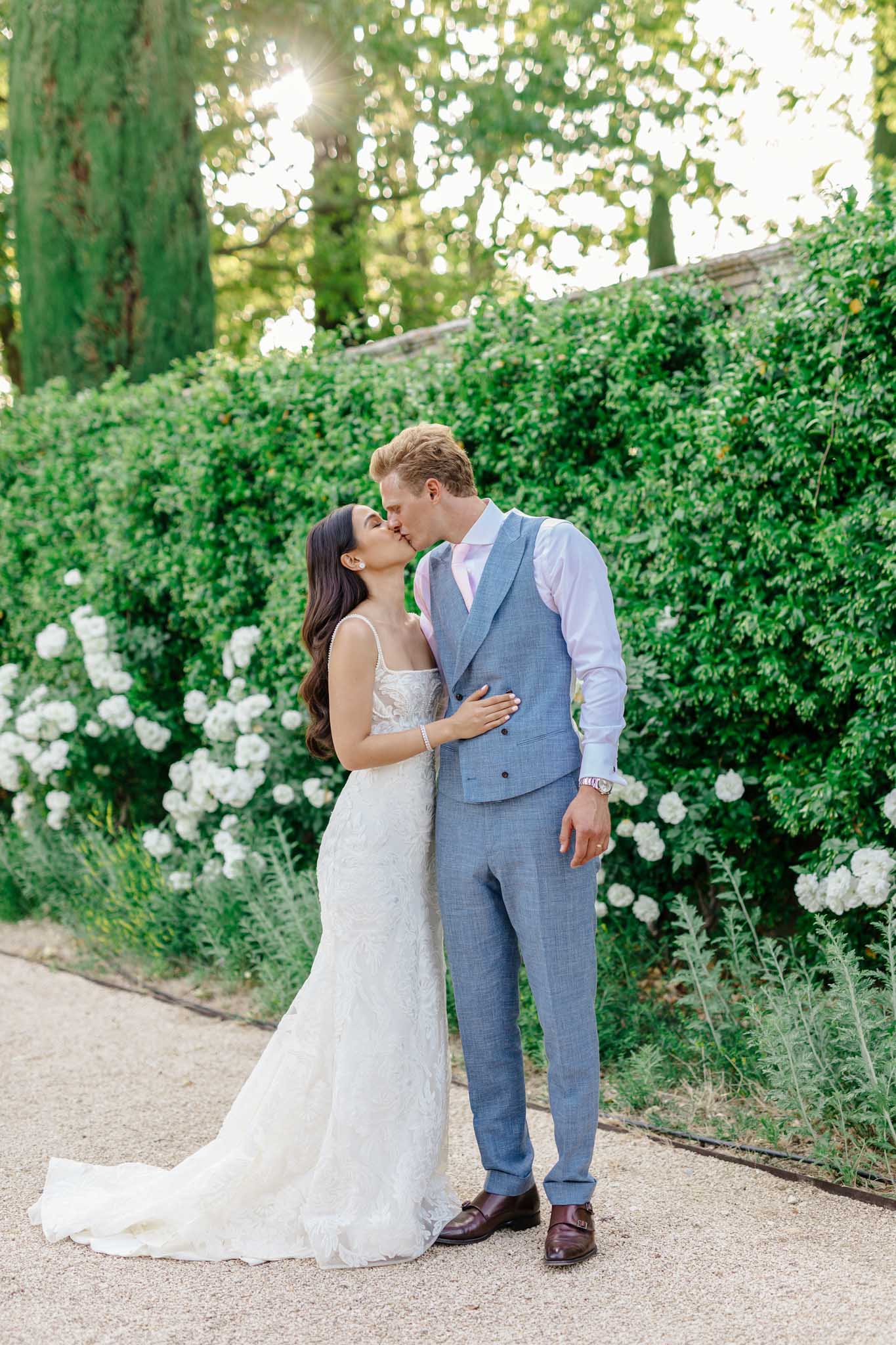 Bride in ivory lace gown and groom in slate blue linen suit kissing in garden courtyard with white flowering shrubs